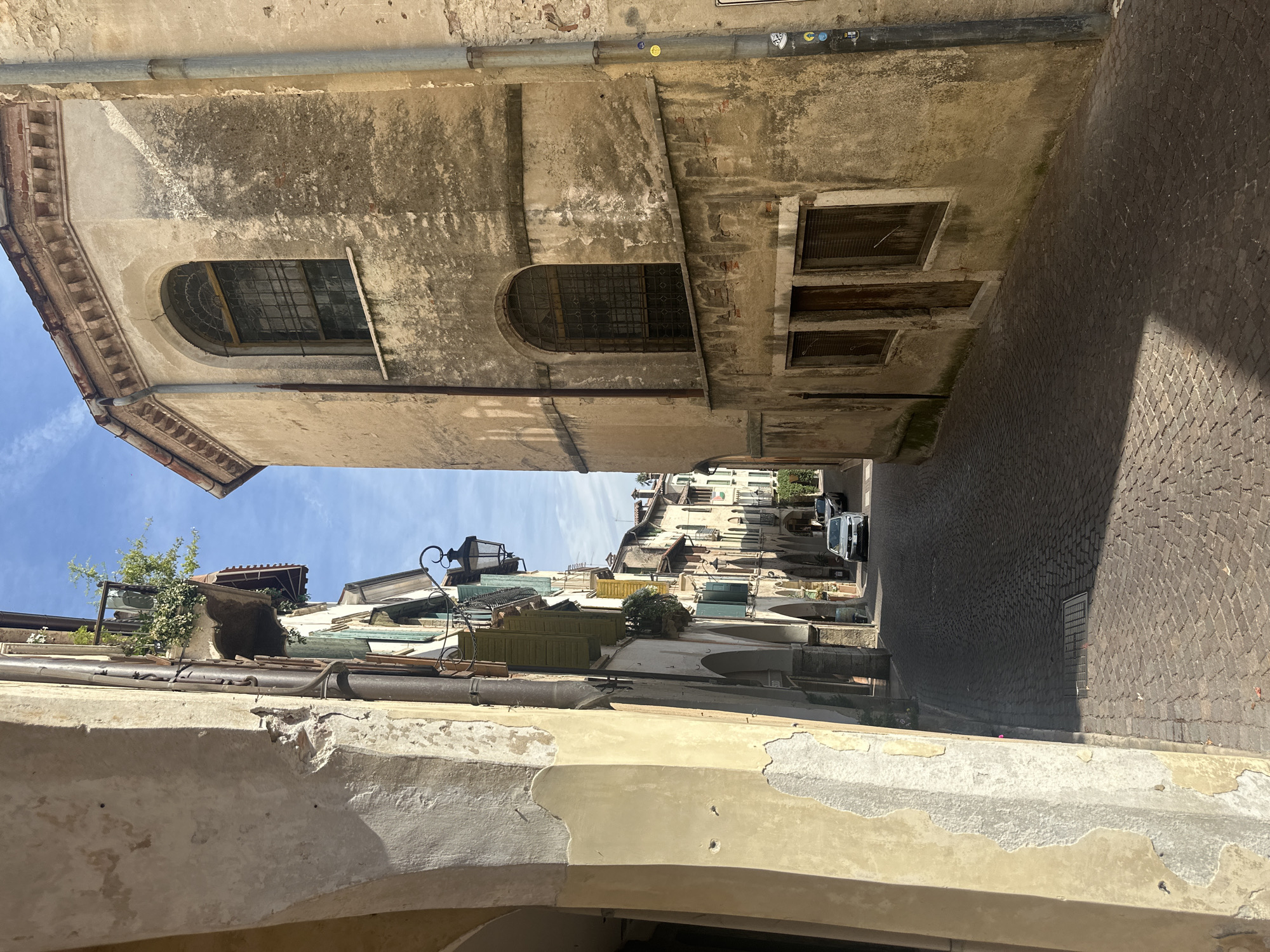 The image depicts a narrow cobblestone street in Maser, Italy, flanked by aged buildings. On the left, a pale yellow building with peeling plaster dominates the foreground, featuring arched openings. To the right, a beige, weathered building with a curved facade showcases arched windows with intricate metal grilles. The street continues into the distance, lined with more buildings displaying architectural details such as balconies with greenery and awnings. Sunlight casts shadows across the cobblestones, indicating a clear day. A white car is parked on the street in the mid-ground, and a dark car is visible further down the street. The sky is blue with scattered clouds.