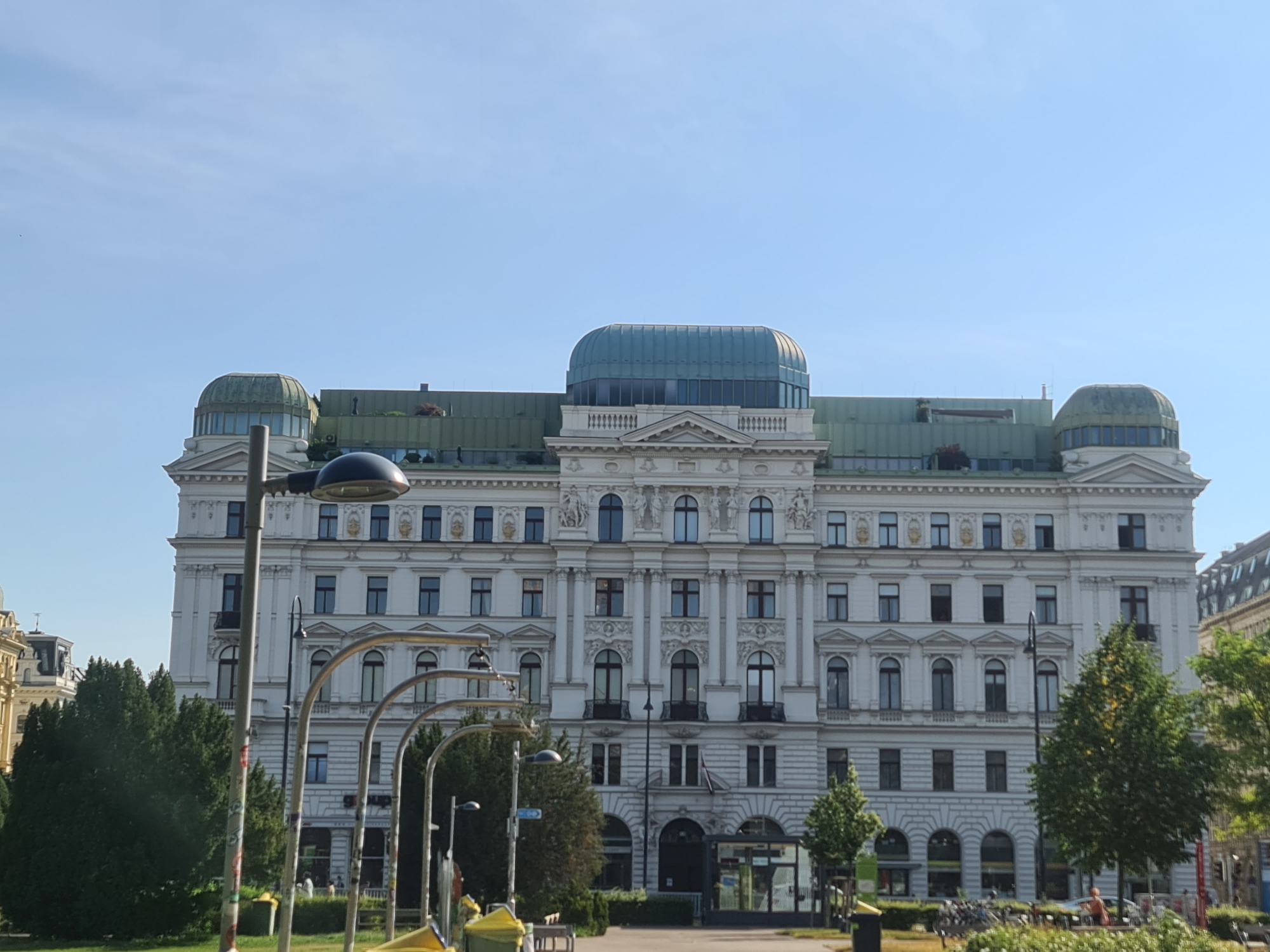 This image shows a stately white building in Vienna, Austria, under a clear blue sky. The building has a grand facade with numerous windows, decorative elements, and two prominent domed structures on either side of a central glass-enclosed section on the roof. In the foreground, there are several street lamps with dark fixtures and curved supports, some of which appear to be decorated with graffiti. Lush green trees and some smaller bushes are visible, as is a paved area with what appear to be street furniture, including benches and trash bins. In the background, glimpses of other buildings and street traffic suggest an urban setting.