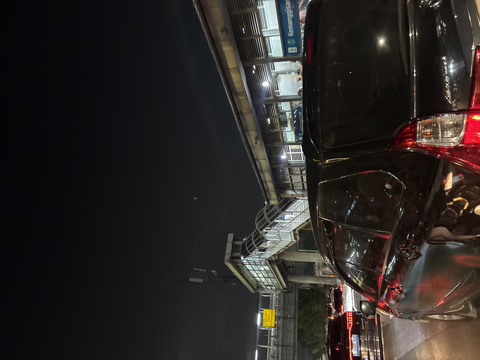 The image depicts a nighttime street scene in West Jakarta, Indonesia, characterized by heavy traffic and illuminated infrastructure. In the foreground, a black vehicle, possibly an SUV or MPV, is prominently displayed, its taillights reflecting red light. To its left, another black car with license plate "B 1403 NDQ" is visible. Further ahead, a white car is also in traffic.
The background features an elevated pedestrian walkway and a bus stop structure. A yellow sign with Indonesian text is mounted on the walkway, likely providing directions or information. The bus stop building has horizontal louvers and displays signage with the word "Kemanggisan" multiple times. Several people are seen on the stairs of the walkway and inside the bus stop structure. The environment is urban, with the presence of tall structures and traffic suggesting a busy city. The overall atmosphere is dark, as it is nighttime, with artificial lights from the infrastructure and vehicles illuminating the scene.