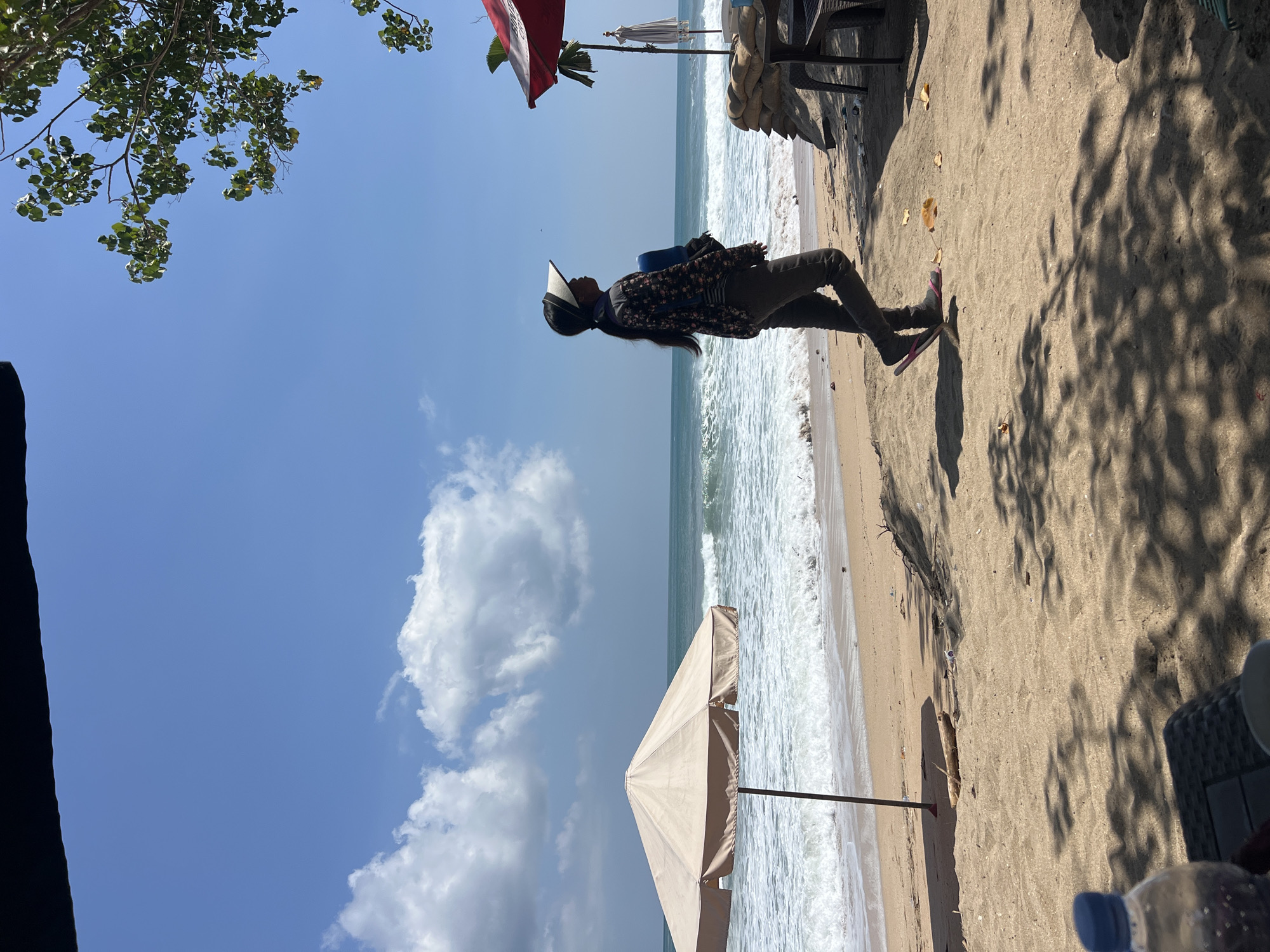 A person walks along a beach in Kuta, Indonesia, carrying a blue object. The person is wearing a patterned long-sleeved shirt, dark pants, a visor, and flip-flops. They are walking from left to right, with their body facing away from the camera and their head turned to the right. The beach is sandy, with a few pieces of driftwood and scattered leaves. The ocean is to the left, with waves breaking on the shore. There are two beach umbrellas, one light beige and one red and white. Sandbags are stacked near the red and white umbrella, which is part of an outdoor seating area with a table and chairs. The sky is blue with scattered white clouds. The image is taken from a low angle, with the shadows of trees and foliage cast on the sand in the foreground. A partially visible water bottle and a dark surface that appears to be a table are in the lower left corner.