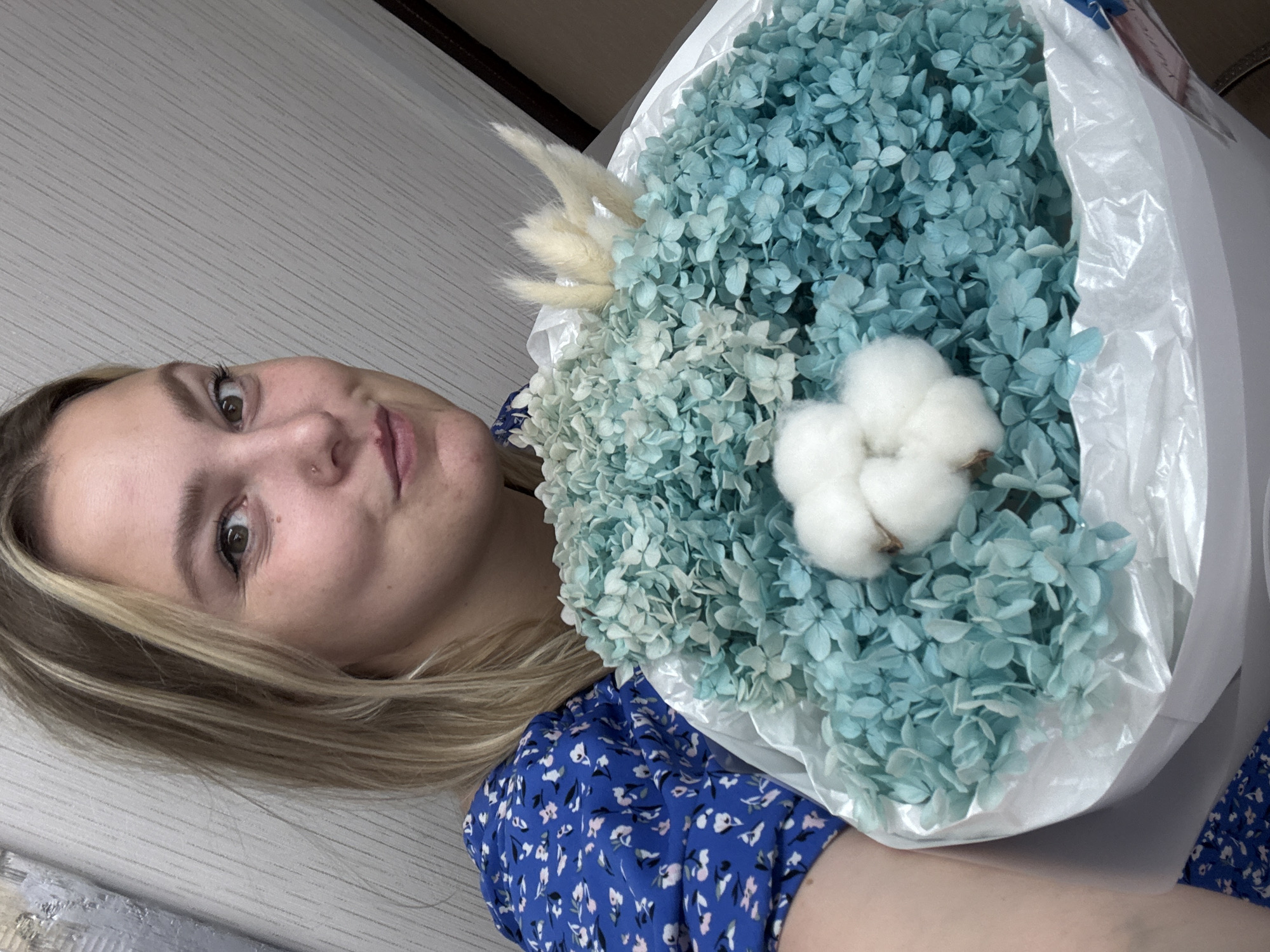 A young woman with blonde hair, wearing a blue floral dress, is holding a large bouquet of dried blue hydrangeas and cotton balls. She is looking at the camera with a slight smile. The bouquet is wrapped in white paper. The background is a light-colored, textured wall. The lighting suggests it is daytime. The overall mood is casual and cheerful. The image appears to be a selfie, with the woman holding the bouquet and phone to capture the photo. There is no visible text in the image. The image is oriented vertically.