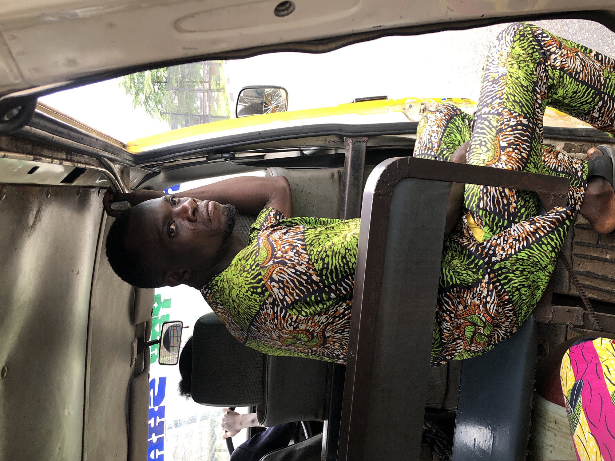 A dark-skinned man wearing a brightly colored green and orange patterned dashiki and matching trousers sits in the passenger seat of a vehicle, likely a shared taxi or minivan, in Nigeria. His left arm rests on the back of the seat as he looks off to the right with a stern expression. The interior of the vehicle is visible, including a rearview mirror mounted to the ceiling, a dark-colored seat behind him, and part of the steering wheel and dashboard. Outside the open door of the vehicle, trees and foliage are visible, suggesting an outdoor urban or suburban setting. The vehicle's interior appears utilitarian and shows signs of wear and tear, consistent with common public transportation in Nigeria. The image captures a moment of transit, highlighting the man's attire and the environment of the vehicle.