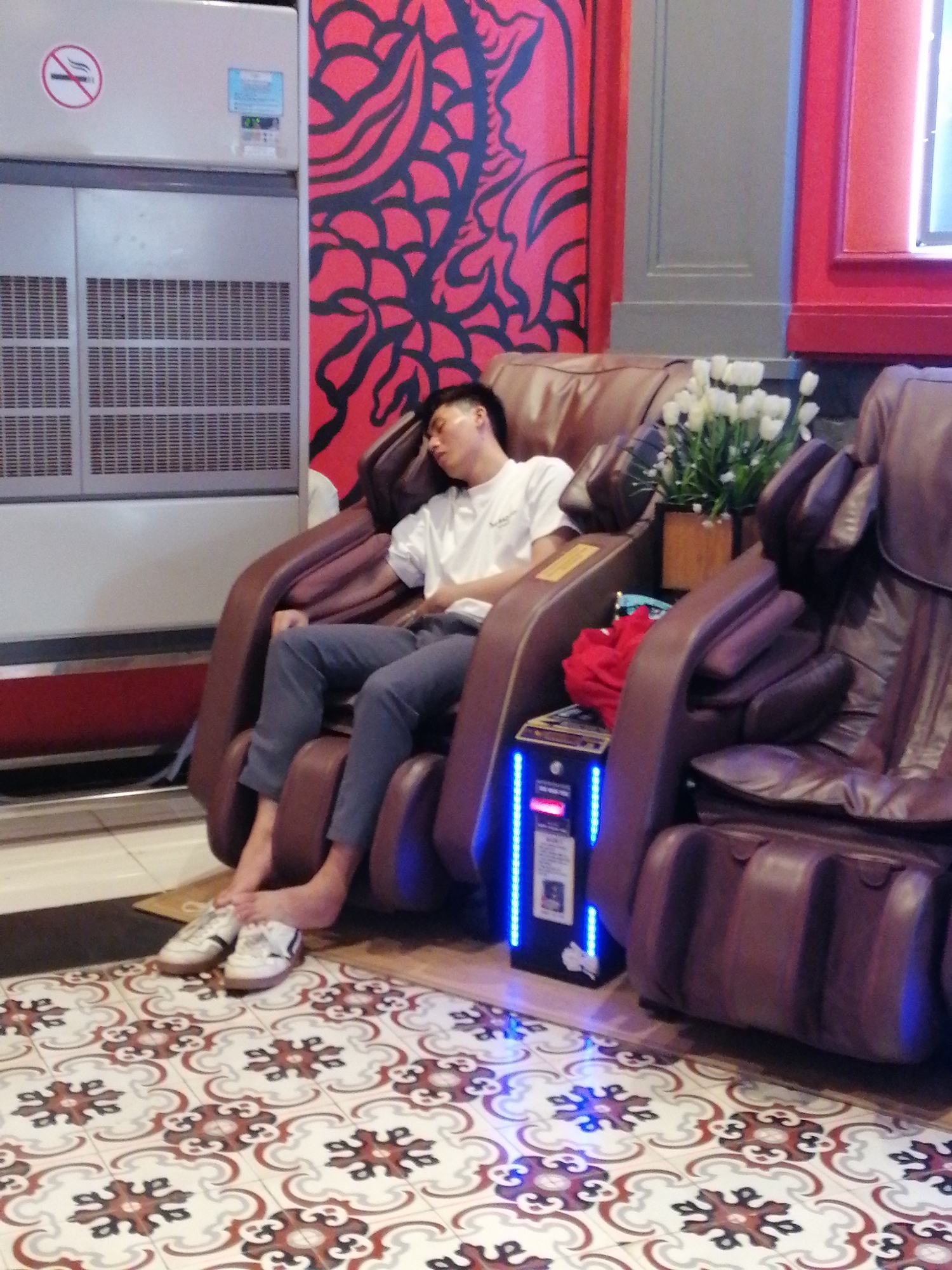 A man is reclined in a brown leather massage chair in a commercial establishment in Bai Chay Ward, Vietnam. He is wearing a white t-shirt and grey pants. His shoes are on the floor near his feet. A second massage chair is beside him, and a small stand with blue lights is next to his chair. A potted plant with white flowers is visible between the chairs. A large, decorative wall mural in red and black is behind the chair. A large, grey air conditioning unit is also visible in the background with a no-smoking sign on it. The floor is tiled with a patterned design in red, brown, and white.