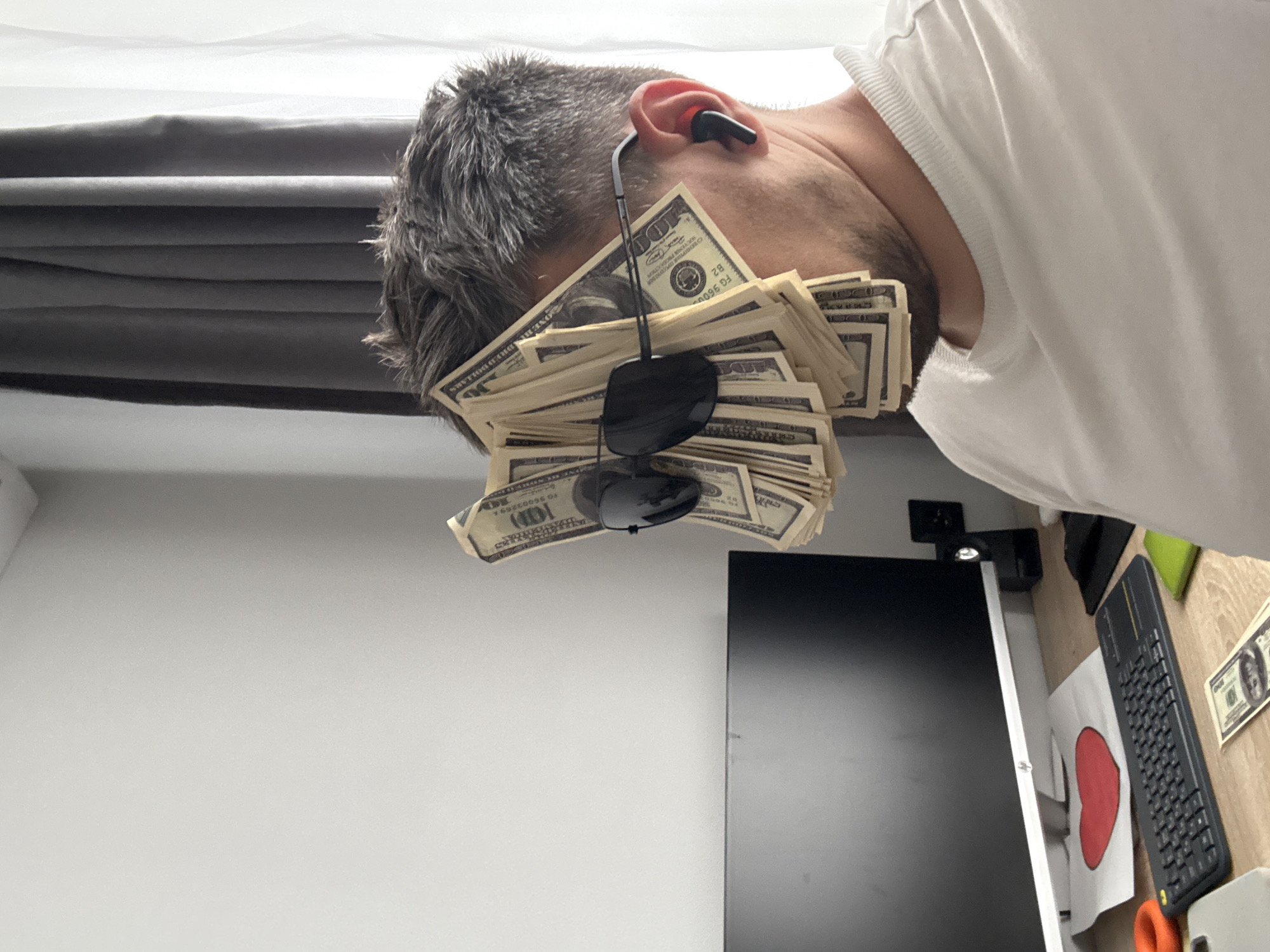 A man with gray hair and a white t-shirt is lying down with a stack of American $100 bills on his face. Sunglasses are resting on the stack of money. The man has a black wireless earbud in his ear. A gray curtain is behind him, and a computer monitor, keyboard, and desk are visible in the background. A $100 bill is also lying on the desk. There is no indication in the image that it was taken in Rostov-on-Don, Russia.