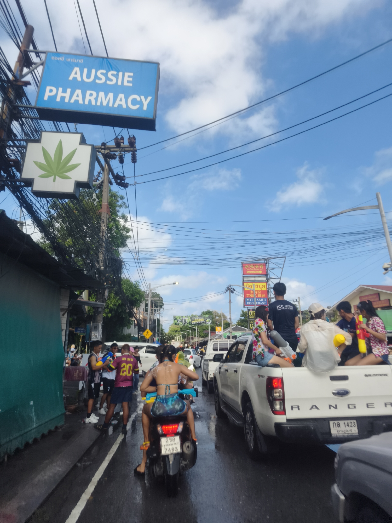 The image depicts a street scene in บ้านบางรักษ์, Thailand, possibly during a water festival. On the left, a sign reads "Aussie Pharmacy" above a green cannabis leaf symbol. The street is wet, and people are gathered on the sidewalk and in the road, some holding water guns. A woman on a motorcycle is driving away from the camera. A white Ford Ranger pickup truck is in the middle of the road, its bed filled with people holding water guns. Other vehicles are visible in the background, and overhead power lines are strung across the sky. Signs are present with writing in Thai. The sky is partly cloudy.