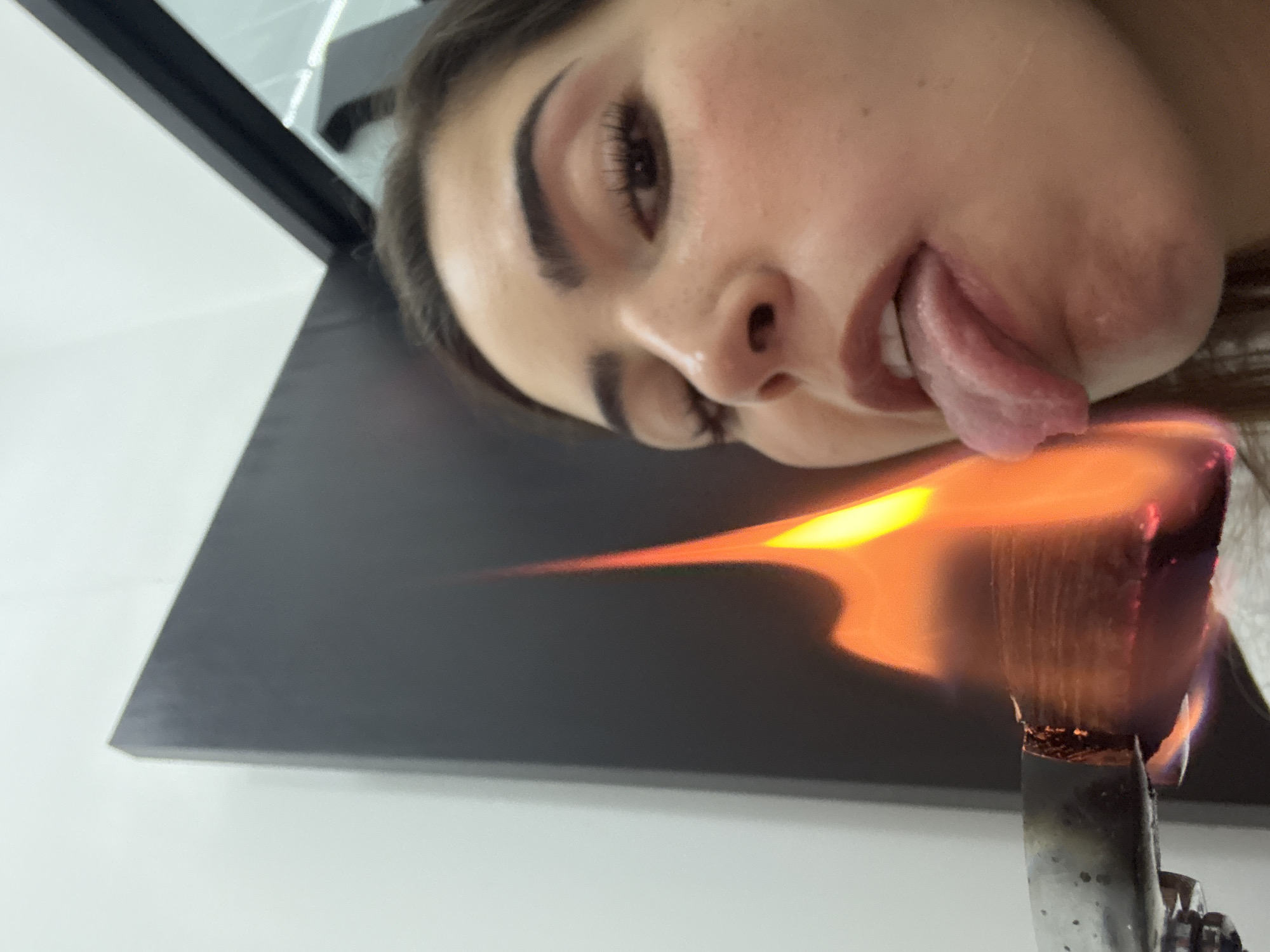 A photograph depicts a person, presumably a young woman, with brown hair, fair skin, dark eyebrows, and eyelashes. She is sticking her tongue out. Beside her, a lit object, possibly a moxa stick, is burning, producing orange flames. The background is a gray surface against a white wall. The location of the image cannot be determined from the provided picture.