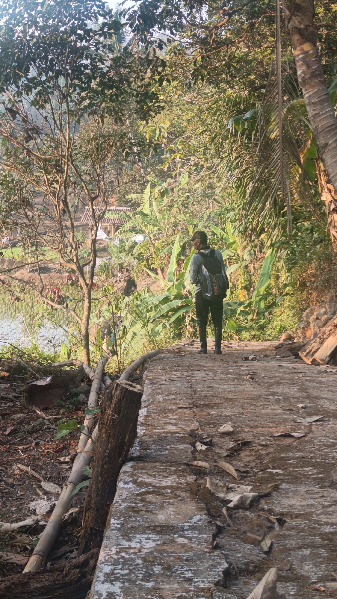 A man stands on a concrete pathway overlooking a body of water in a rural setting, possibly in Sumedang, Indonesia. The man is wearing a grey shirt, dark pants, and black boots, with a backpack on. The pathway is cracked and shows signs of wear. Lush green vegetation, including trees and banana plants, surrounds the area, with some structures visible in the distance. The sun is casting long shadows, indicating either early morning or late afternoon.