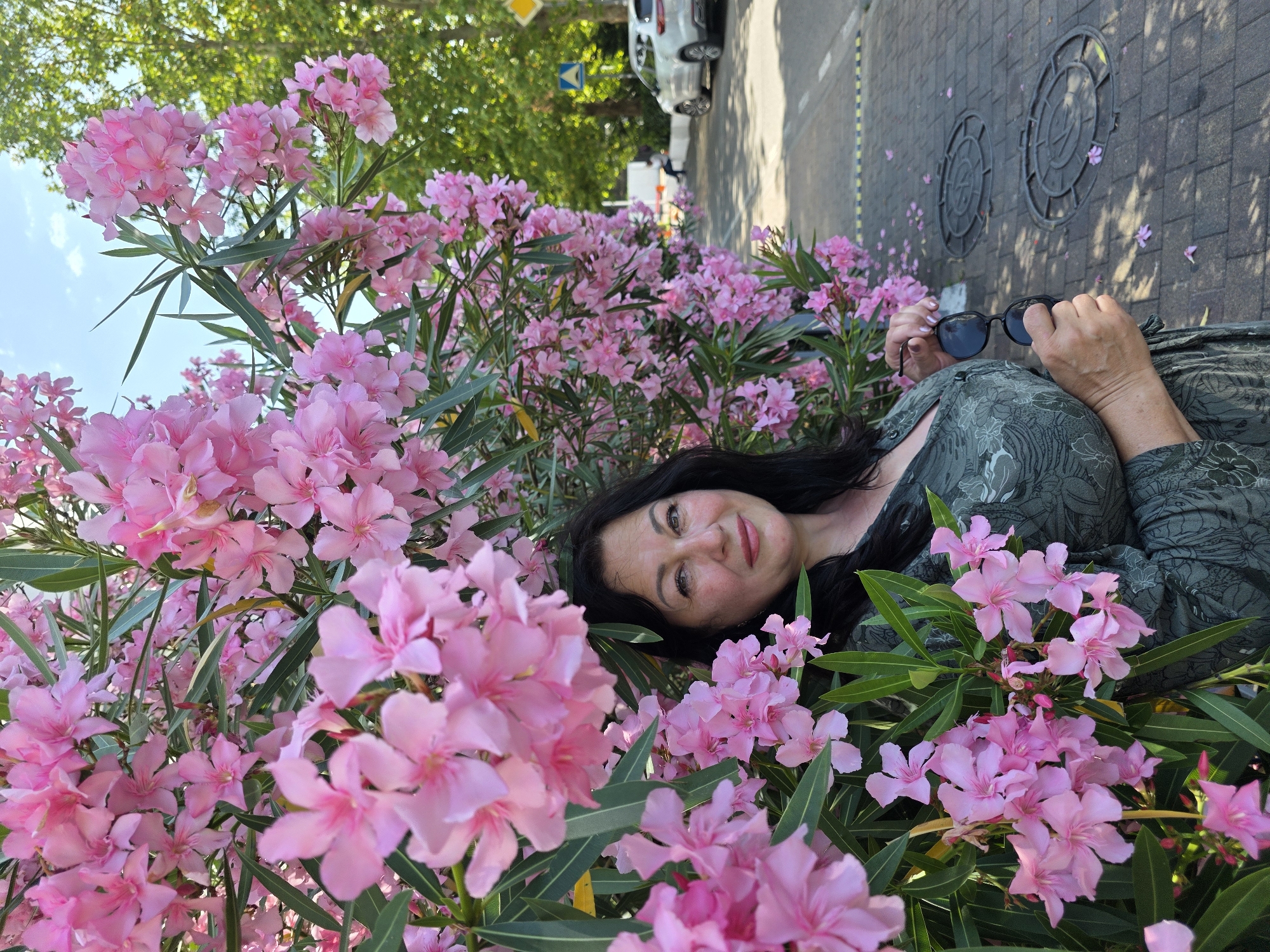 A medium shot shows a woman with dark hair and olive skin standing in front of a wall of pink oleander flowers. She is wearing a green patterned dress and holding a pair of sunglasses in her right hand. The woman is smiling and looking towards the camera. Behind her, a street is visible with a car, road signs, and pedestrian crossing. The scene is set outdoors on a sunny day. The location is identified as Sochi, Russia.