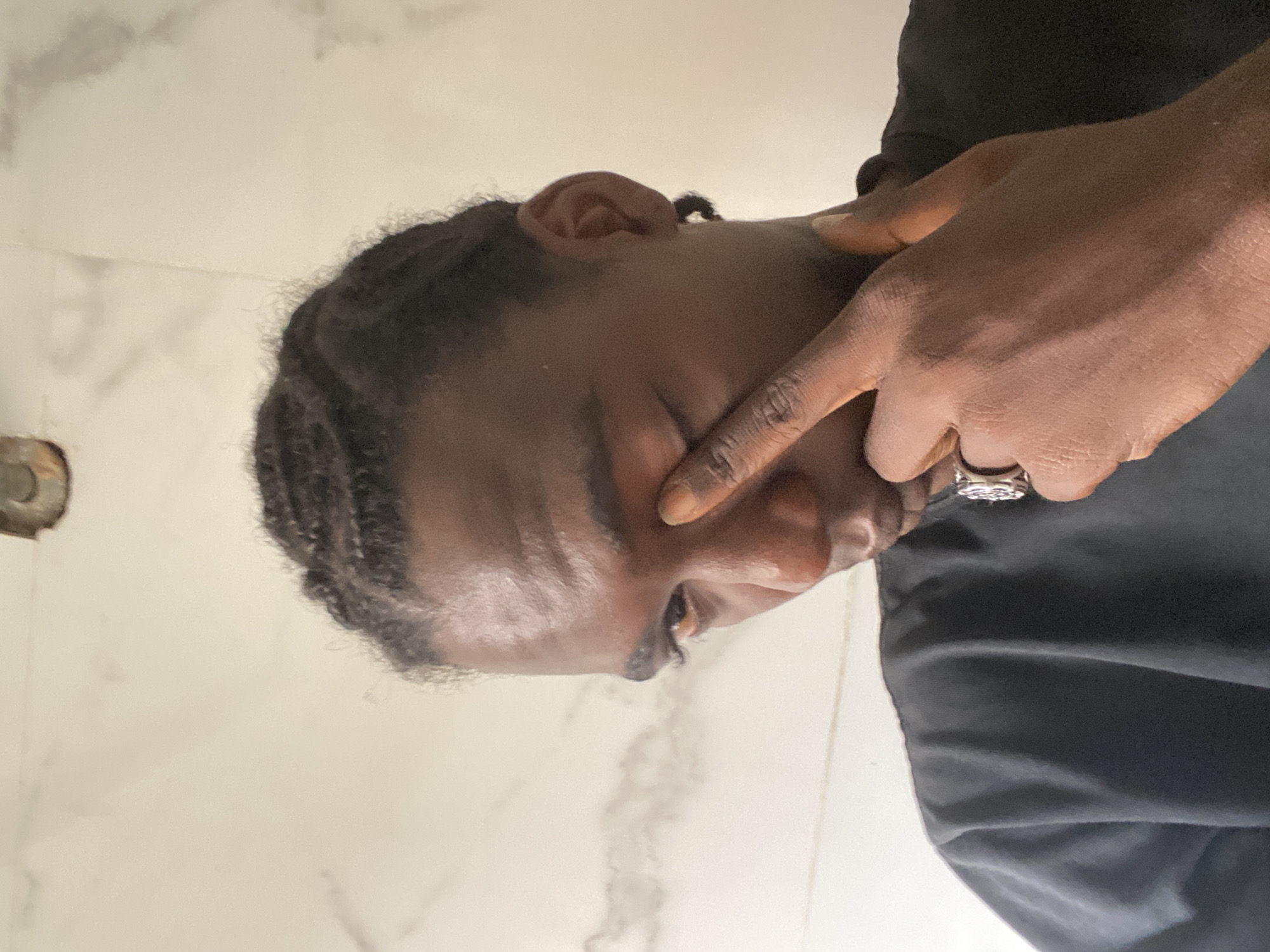 A person of African descent with braided hair and brown skin, wearing a dark-colored shirt, is seen in a close-up, side-profile shot. Their right hand, adorned with a ring on the ring finger, is touching their face near the eye. The background is a white surface, possibly a tiled wall. The location is likely a bathroom. The setting implies an indoor, personal space. Given the prompt, it is suggested that this is taken in Lagos, Nigeria.