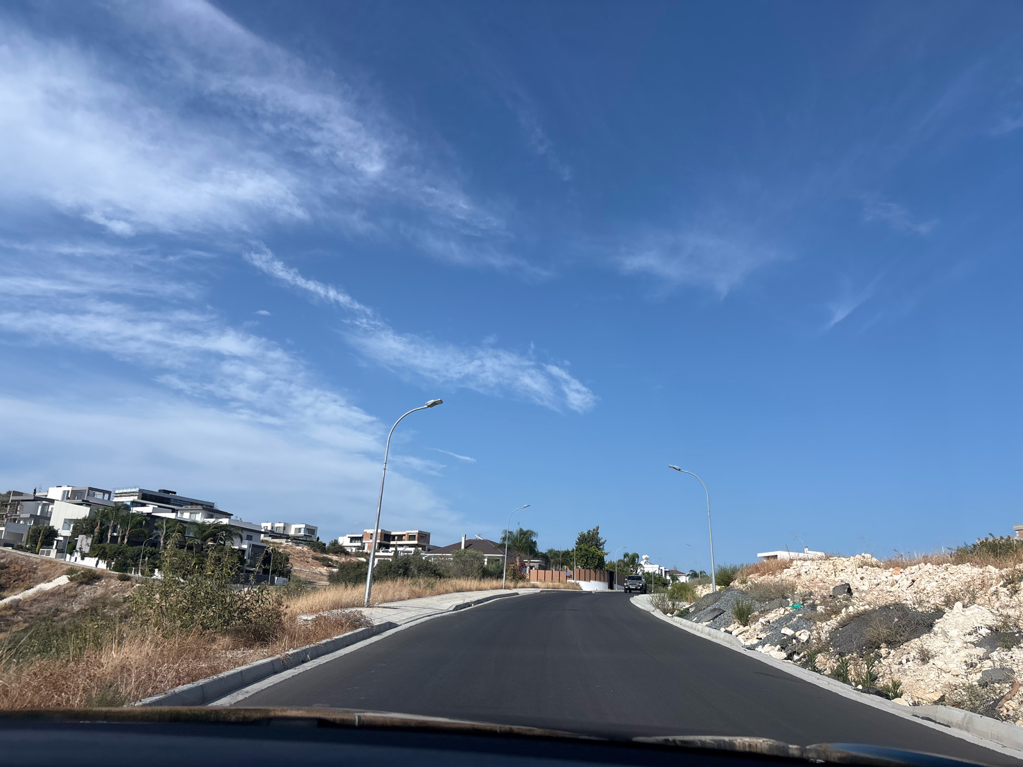 A road curves uphill through a residential area on a sunny day. Modern houses with flat roofs and white facades are situated on the slopes to the left, with some construction visible. Dry, yellowish grass and sparse green shrubs cover the hillsides. Two streetlights with modern designs are positioned along the road. In the distance, a dark-colored SUV is visible on the road. The sky is a clear, bright blue with wispy white clouds. The scene appears to be in Agios Athanasios Municipality, Cyprus, given the architecture and landscape.