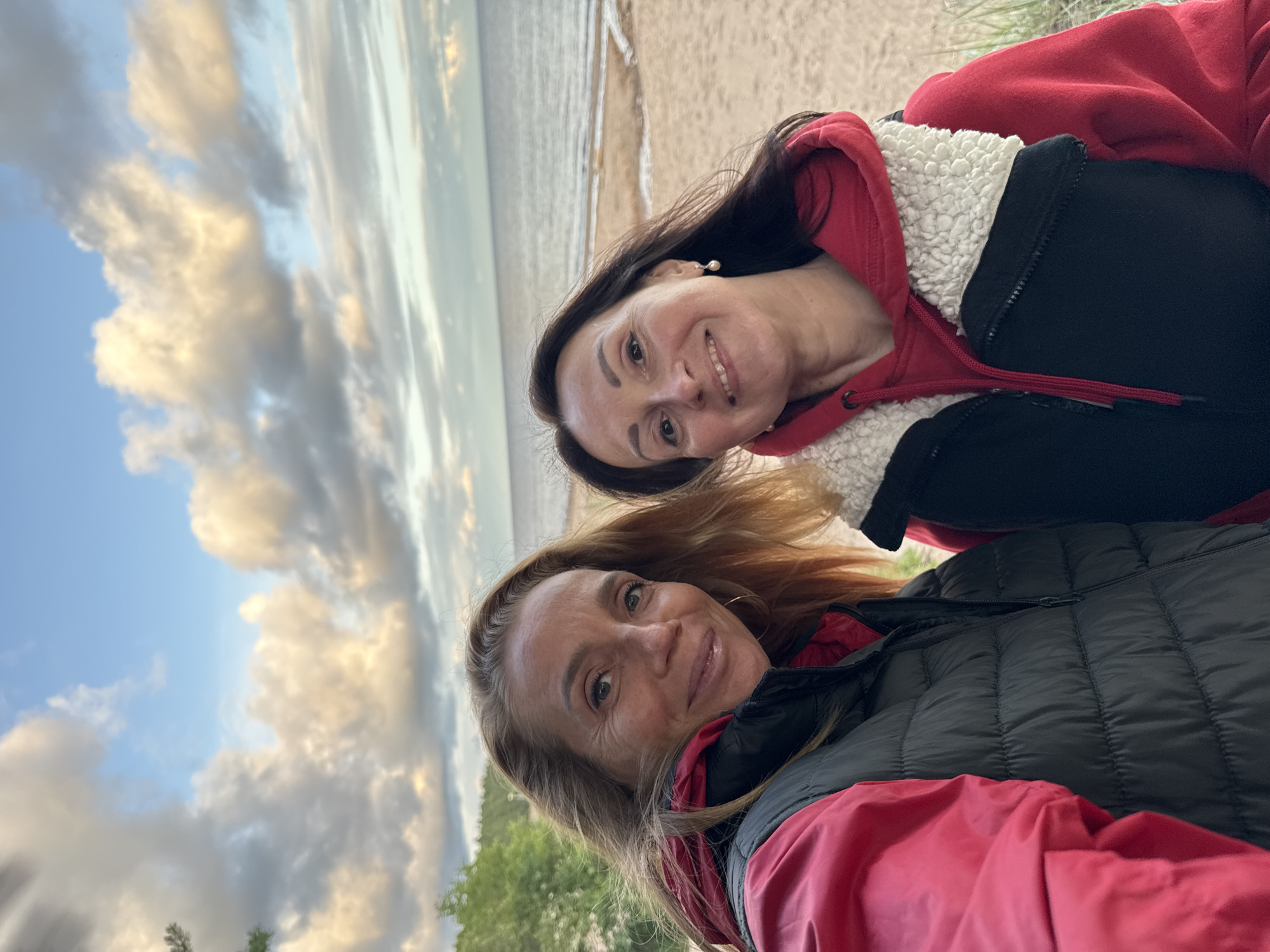 Two women are standing on a beach with the sea and sky in the background. The woman on the left has blonde hair and is wearing a red jacket and a dark vest. The woman on the right has dark hair and is wearing a red hooded sweatshirt and a dark vest with a white fuzzy collar. They are both smiling at the camera. The beach is sandy with some sparse grass. The sky is cloudy with the sun setting. The location is Zelenaya Roshcha, Russia.