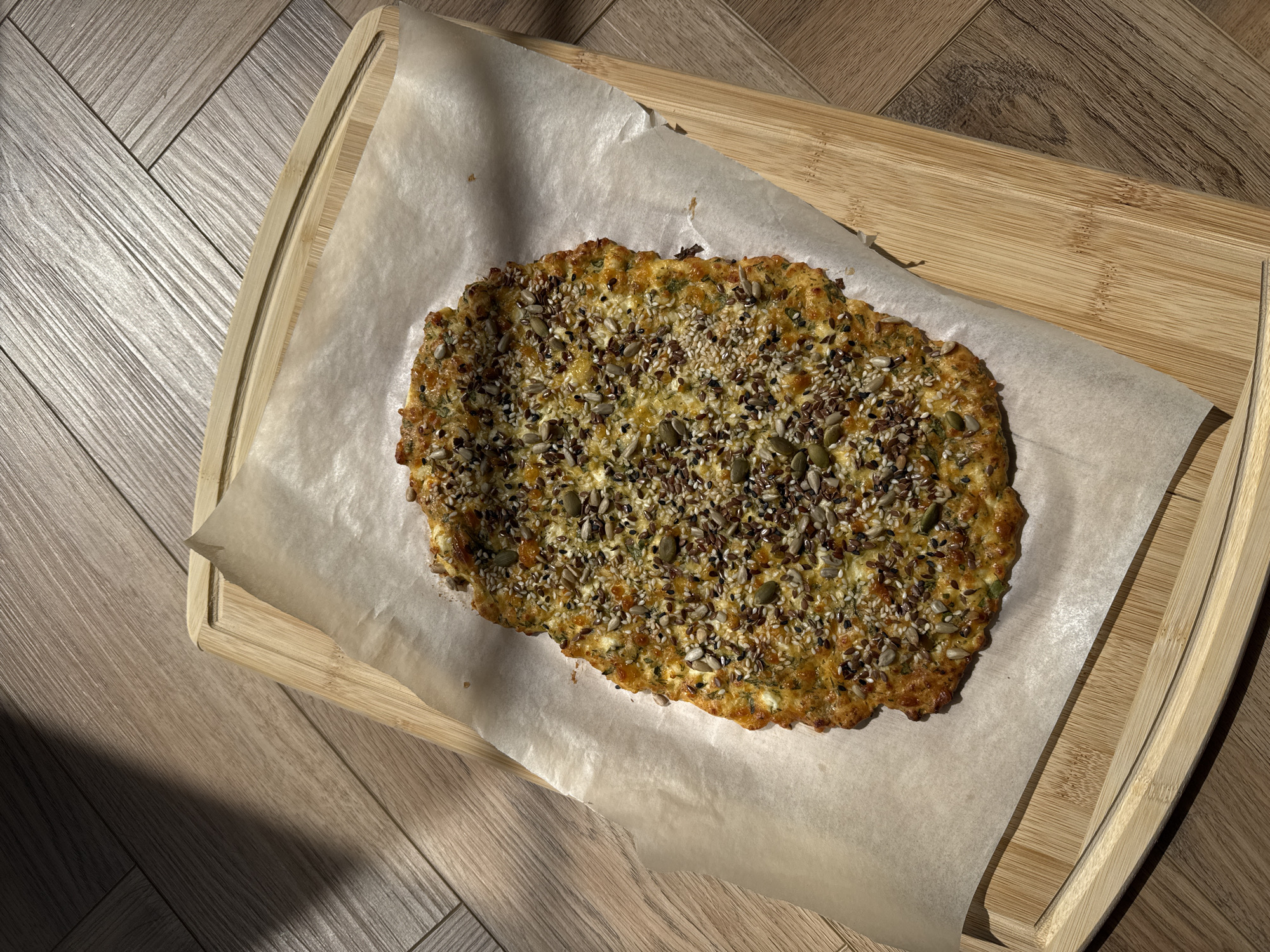 Here is a top-down shot of a rectangular, light-colored wooden cutting board placed on a wood-grain patterned surface, likely a floor or table. A sheet of parchment paper lies on the cutting board. The parchment paper holds a seed-covered baked item, possibly a bread or flatbread. The baked item has an irregular, oval shape and is covered with various seeds, including pumpkin seeds, sesame seeds, and flax seeds. Strong natural light illuminates the scene, creating distinct shadows. The location appears to be indoors, but the specific city and region are unknown.
