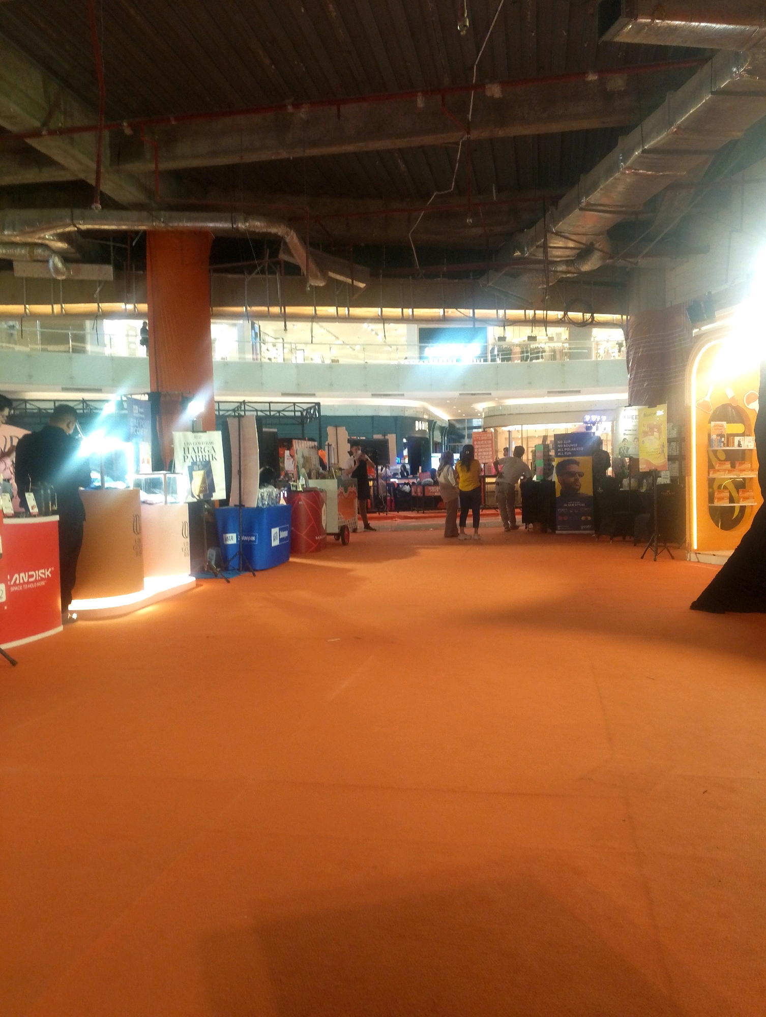 This image depicts an indoor event, likely a product launch or exhibition, set within a spacious, modern shopping mall. The environment features an orange carpet covering the floor, with various booths and displays arranged around. In the background, the mall's upper levels and ceiling structure, including exposed pipes and ventilation systems, are visible. Several individuals are present, some interacting with the displays, while others appear to be browsing or working at the booths. One prominent display on the left shows a red panel with the "SANDISK" logo. Another display features a tall poster with the text "WARGA FABRIK" and an image of a gold bar. The lighting is a mix of ambient mall light and spotlights focused on the exhibition areas.