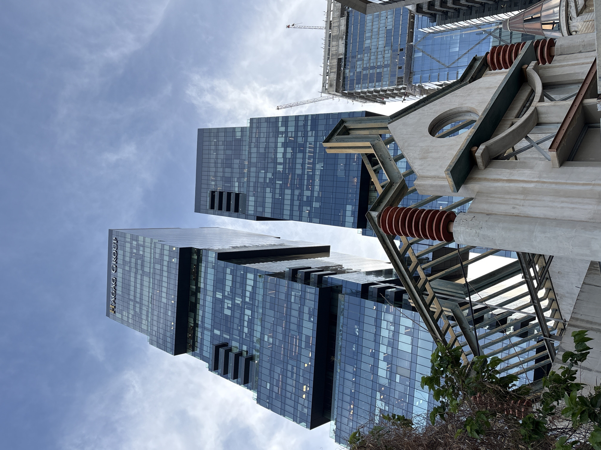 This image shows a low-angle view of modern architecture in Tel Aviv, Israel. Two tall, glass-clad skyscrapers dominate the frame. The building on the left displays the logo "HAGAG GROUP" near its top. A building under construction, with a crane visible, stands further back. A portion of an abstract steel and concrete structure occupies the foreground, with circular cutouts and reddish-brown insulators. Bare trees and a cloudy sky complete the setting.