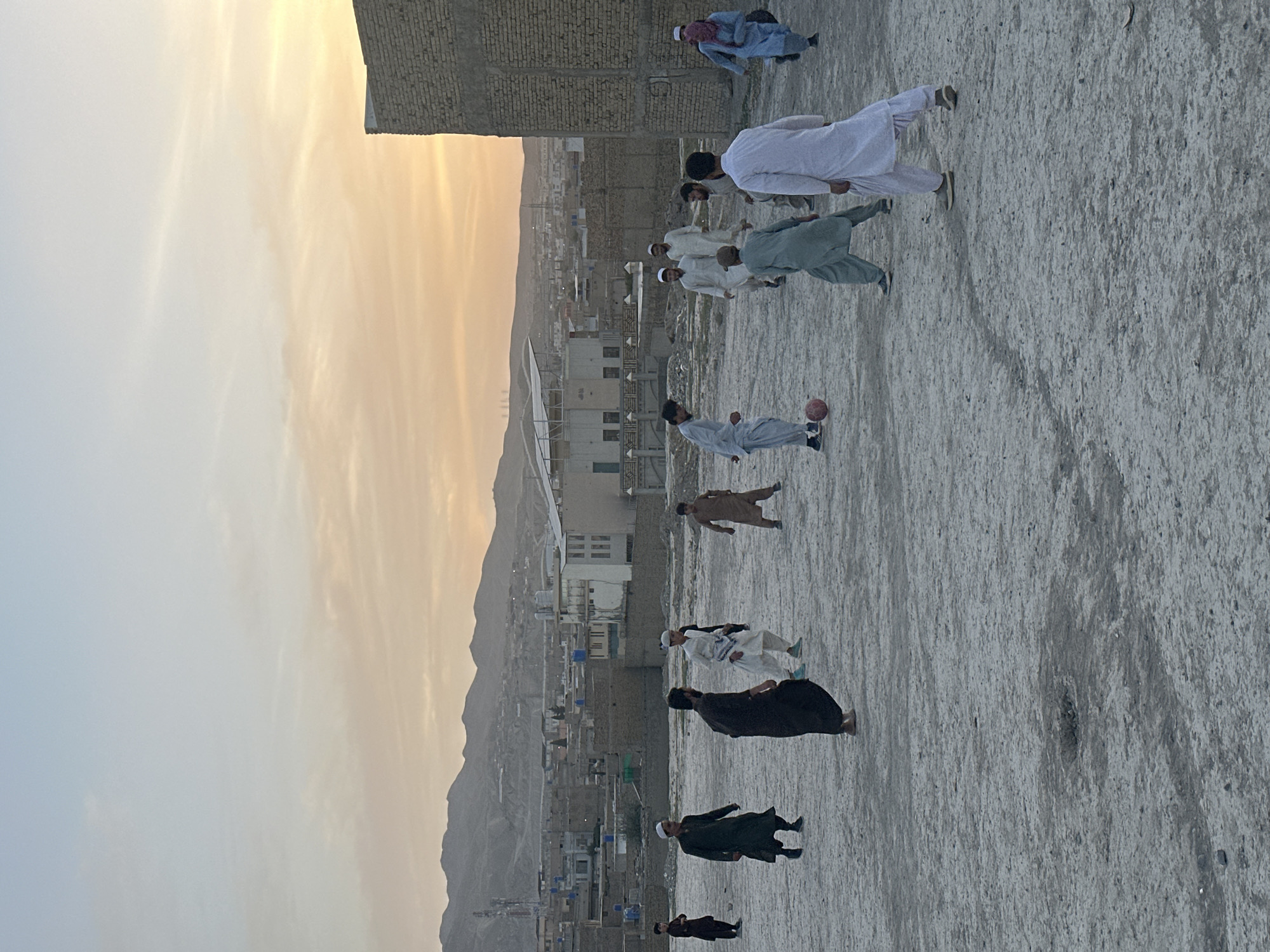 The image shows a group of people playing soccer on a rooftop in Quetta City Tehsil, Pakistan, at sunset. The backdrop includes a cityscape and mountains. The players are wearing a mix of traditional clothing. A soccer ball is visible near one player.