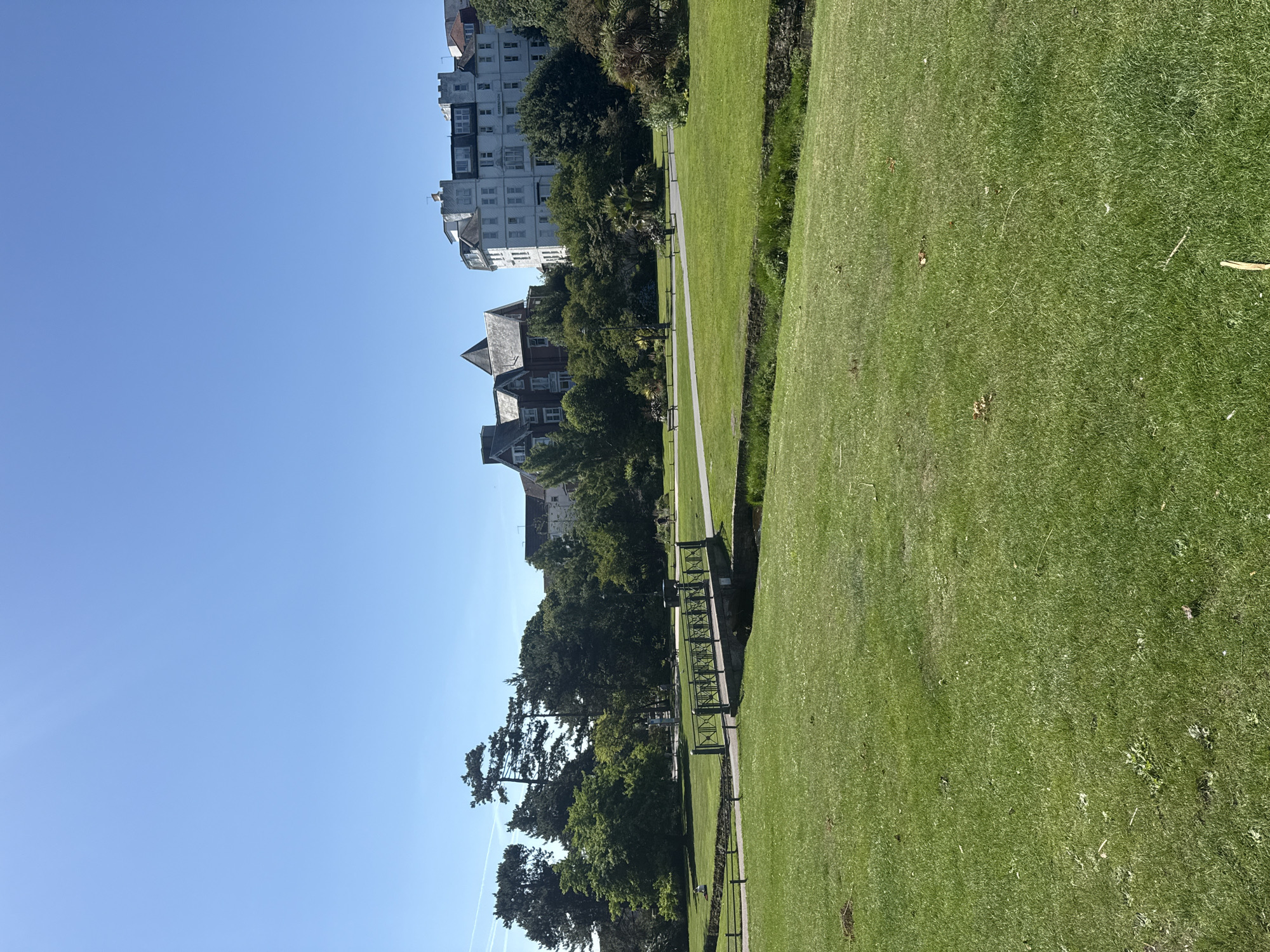 The image depicts a sunny day in Bournemouth's Lower Gardens, a public park in Bournemouth, United Kingdom. In the foreground, a lush green lawn slopes gently upwards. A narrow, stone-arched bridge with ornate metal railings crosses a small stream or ditch that bisects the landscape. Beyond the bridge, a paved pathway winds through the park, bordered by more manicured lawns and mature trees. In the mid-ground, several people are visible: one individual sits on the grass near the bridge, while others walk along the pathway. In the background, large, multi-story buildings with white facades and traditional architecture are visible, suggesting the proximity of the town center to the park. The sky is a clear, bright blue, indicating pleasant weather. The overall scene conveys a peaceful and well-maintained urban park setting.