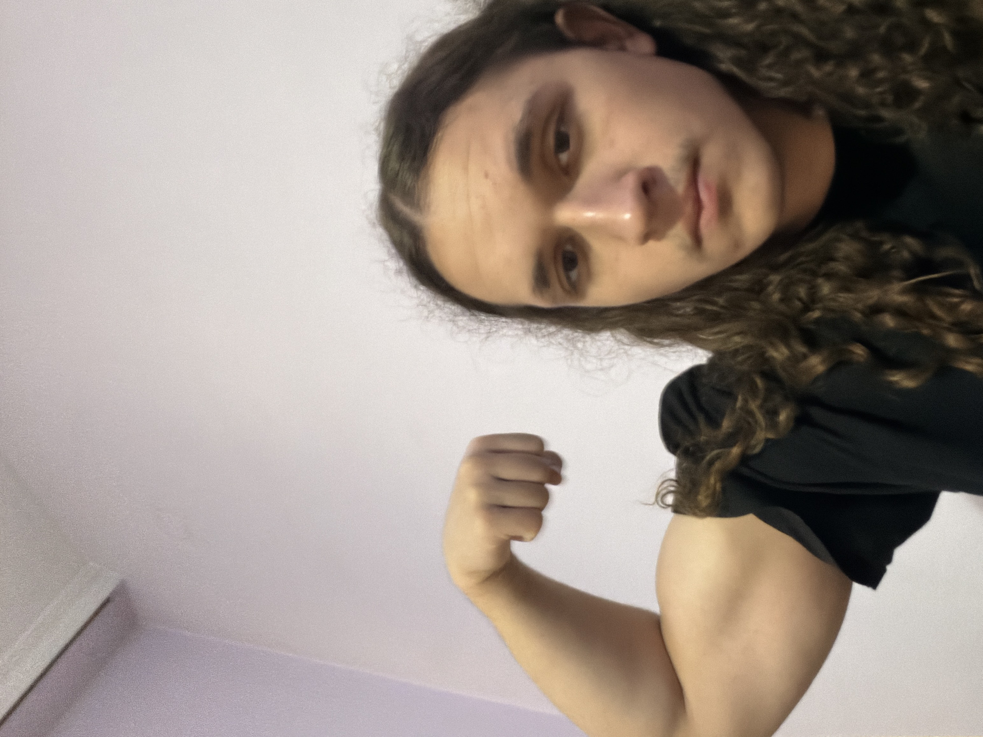 A person with long, curly brown hair and fair skin is posing for a photo. The camera is positioned at a low angle, looking up at the subject. The person is wearing a black shirt and flexing their right bicep. The background consists of a white wall and a hint of a light purple-colored wall in the lower left corner. The location is unspecified, but the prompt specifies that it is in the city of Asenovgrad, Bulgaria. No specific objects, actions, interactions or further environmental details are visible.