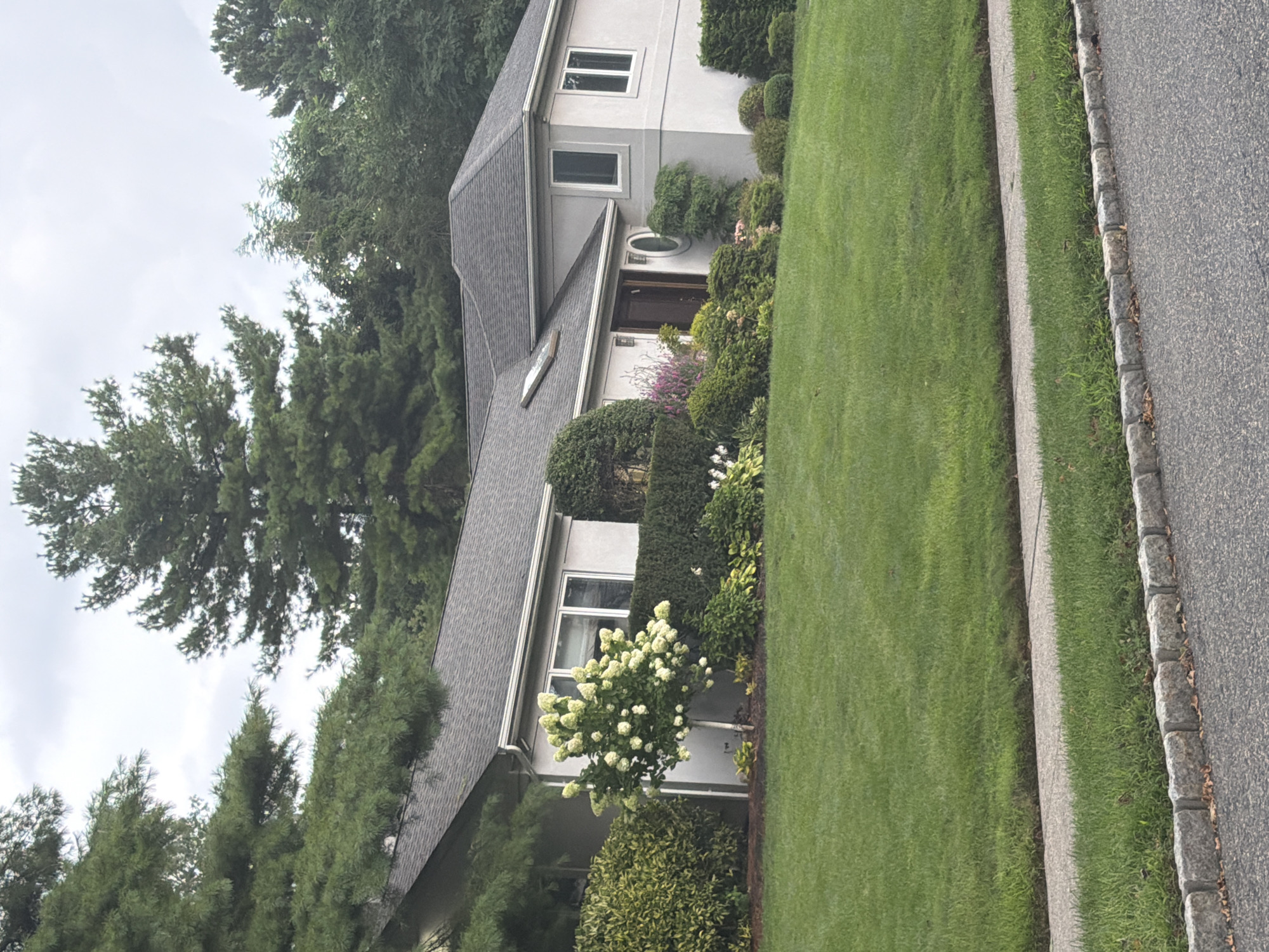 The image depicts a two-story house with a gray stucco exterior and a dark gray asphalt shingle roof. The house is situated on a well-manicured lawn in the Village of Great Neck, United States of America. Tall evergreen trees surround the property, and there is a neatly trimmed hedge in front of the house. A small white flowering tree stands in the front yard, to the left of the entrance. The entrance features a dark wooden door and a round decorative window. The house has several windows, including a large bay window on the side. The foreground shows a paved driveway and a sidewalk with a stone border. The sky is overcast, suggesting a cloudy day. There are no people visible in the image.
