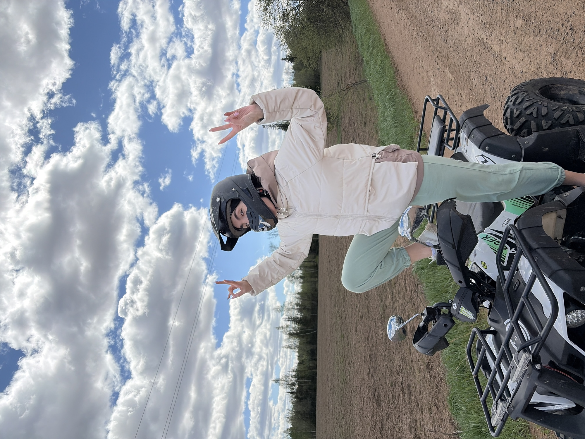 A person wearing a helmet, a beige jacket, and light green pants stands next to a white and black all-terrain vehicle (ATV) in an outdoor setting, with a partly cloudy sky overhead. The ground appears to be a mixture of dirt and grass, suggesting a rural or semi-rural environment. A line of trees is visible in the background. Electrical wires stretch across the sky in the distance. The individual is making a hand gesture, possibly a peace sign or another form of greeting. The exact location is unknown, and it cannot be confirmed that this image was taken in the city of Mozhga, Russia without more context or identifying features.