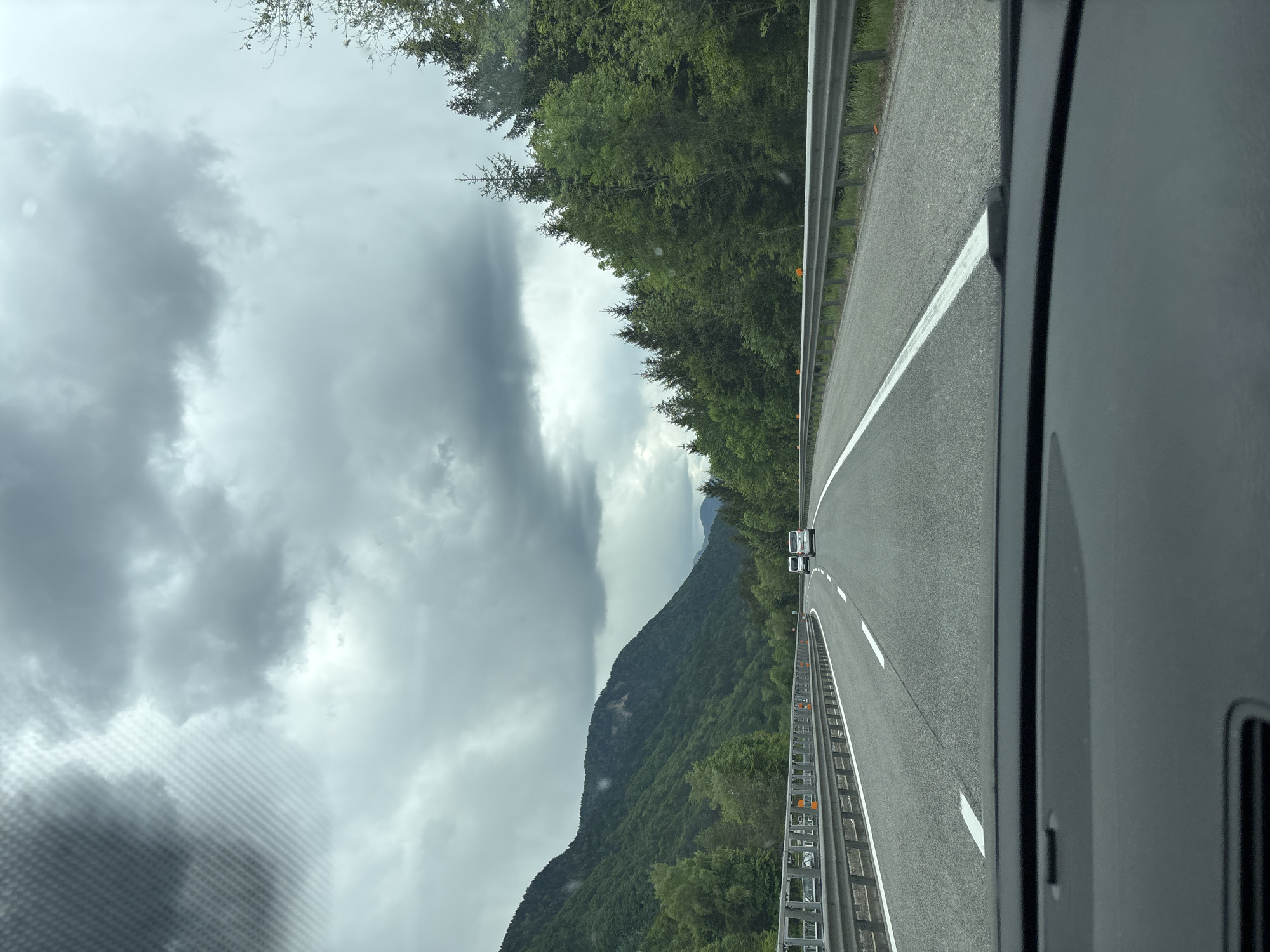 The image shows a view from inside a vehicle traveling on a highway in what appears to be a mountainous region near Malborghetto-Valbruna, Italy. The highway has two lanes separated by a white line, with metal guardrails on both sides. Trees and a mountain are visible on the left, and more trees line the right side of the highway. The sky is overcast with heavy clouds. A vehicle is traveling ahead in the same lane. The interior of the vehicle is visible in the lower right, including part of the dashboard and an air vent.