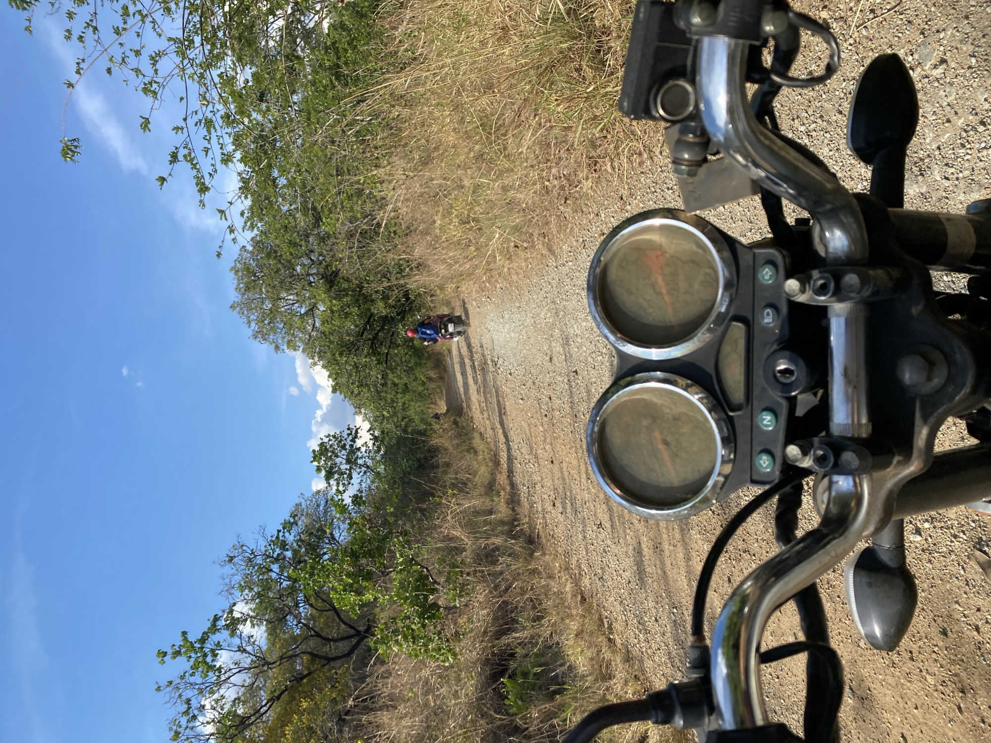 The image shows a motorcycle's handlebar and instrument panel, with two round gauges visible. The view is from the perspective of the motorcycle rider, looking down a dirt road. Another motorcycle, ridden by a person wearing a blue shirt and a red helmet, is visible in the distance on the road. The road is flanked by dry grass and trees. The sky is blue with some clouds. The image was taken in Tinaquillo, Venezuela.