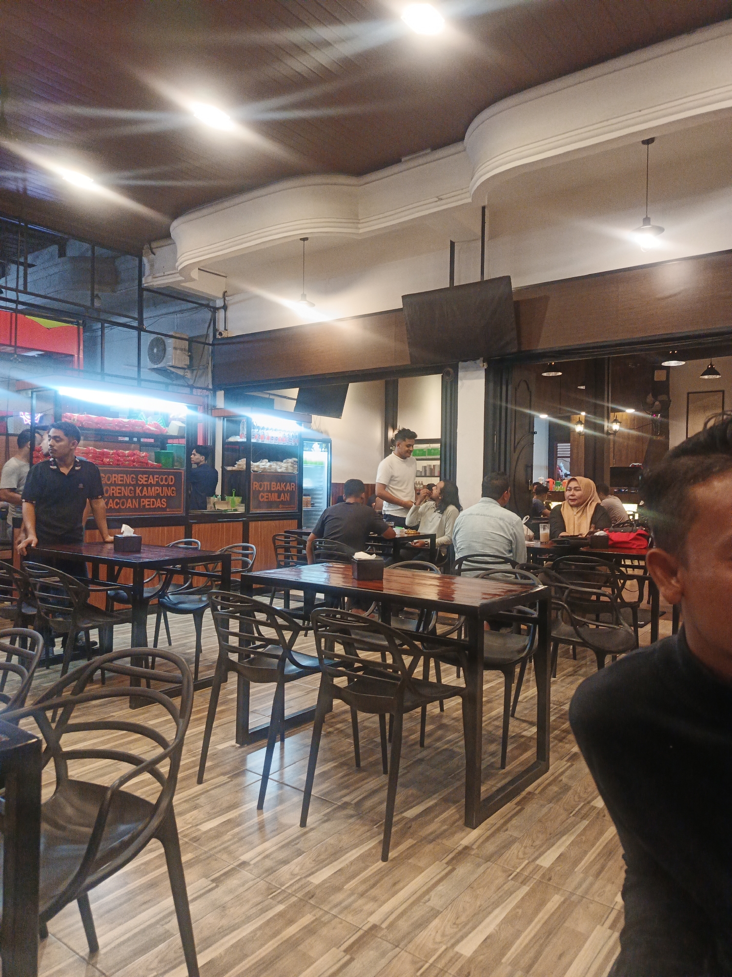 The image depicts the interior of a restaurant in Pantegajah, Indonesia. The dining area features numerous tables and chairs, with a variety of patrons engaged in dining and conversation. The walls are decorated with wooden panels and display menu items, including "GORENG SEAFOOD," "GORENG KAMPUNG," and "ROTI BAKAR CEMILAN." The lighting is warm and ambient, emanating from overhead fixtures. In the foreground, a man in a black shirt stands near a table, while other individuals are seated at various tables throughout the restaurant. The overall atmosphere suggests a casual and bustling dining experience.