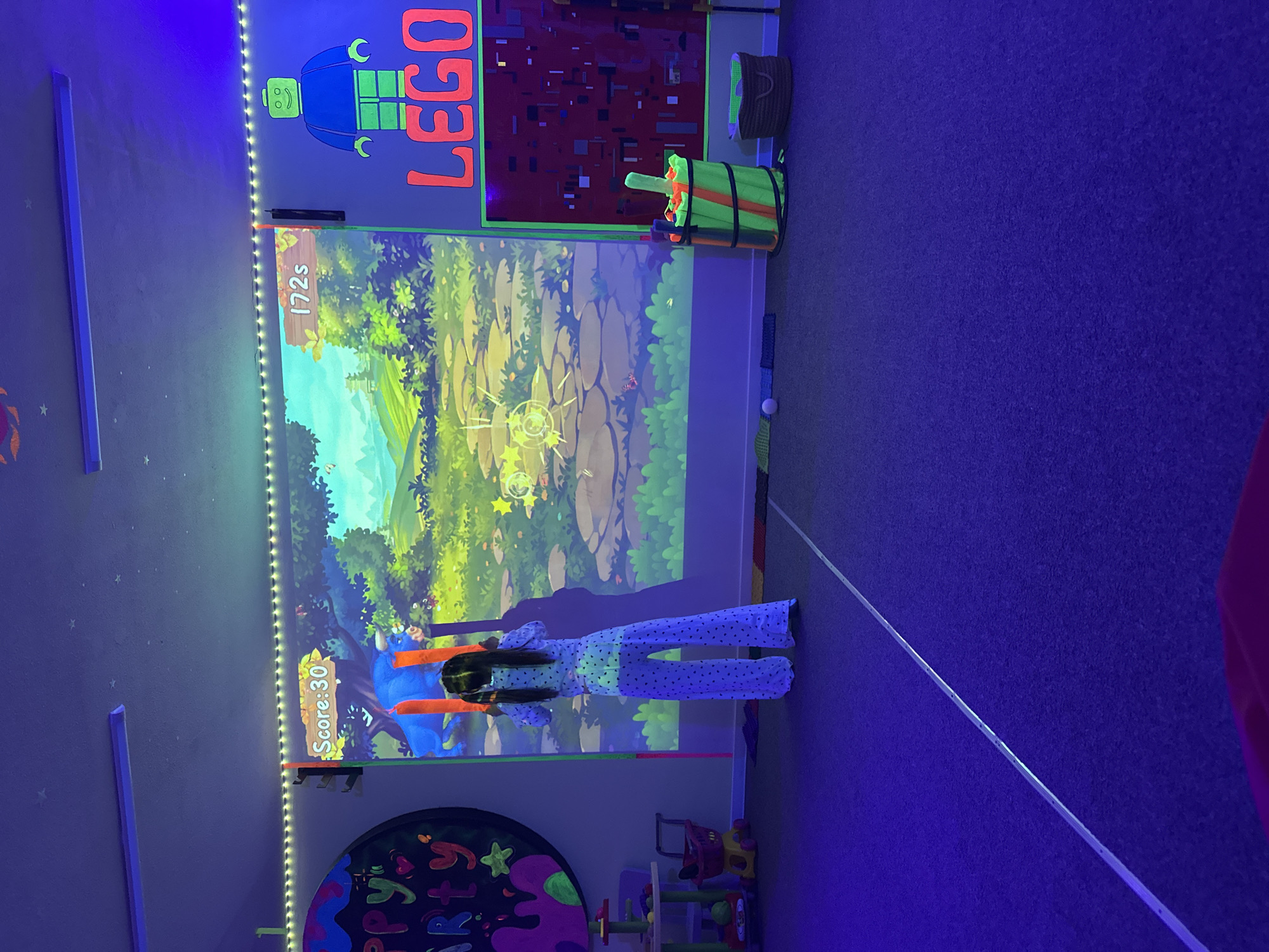 A brightly lit entertainment room features a child playing a game projected onto a large screen. The room is illuminated with blue-toned lighting, and decorations include LEGO-themed designs on the walls, such as a LEGO figure and the word "LEGO" in large letters. The game screen shows a cartoonish landscape with a scoring system.
A child wearing a light-colored, polka-dotted outfit stands in front of the screen, holding a toy blaster. To the side of the screen, there is a bucket filled with colorful objects. The floor is carpeted in a dark color.
Additional room elements include a decorative board with vibrant colors, a small child's cart, and additional lighting details.