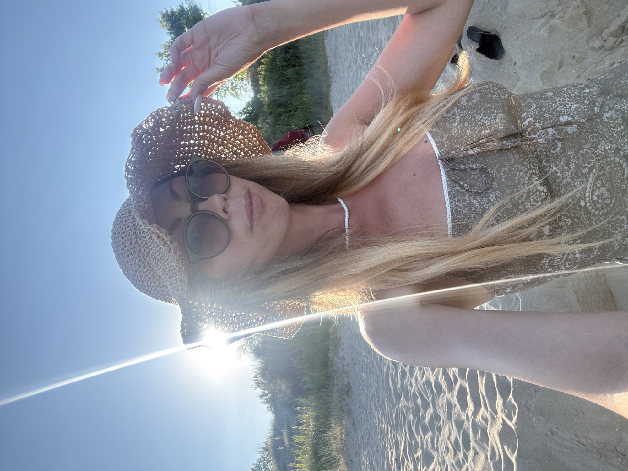 A woman with blonde hair, wearing a straw hat and sunglasses, poses for a selfie on a sandy beach. The sun is shining brightly, casting a lens flare across the image. She is wearing a light-colored dress with a floral pattern and a white beaded necklace. Behind her, there is sand, low vegetation, and a glimpse of trees. A pair of black sandals is visible in the sand to her right. The scene appears to be a sunny day at the beach, possibly in Nesebar, Bulgaria, given the context.