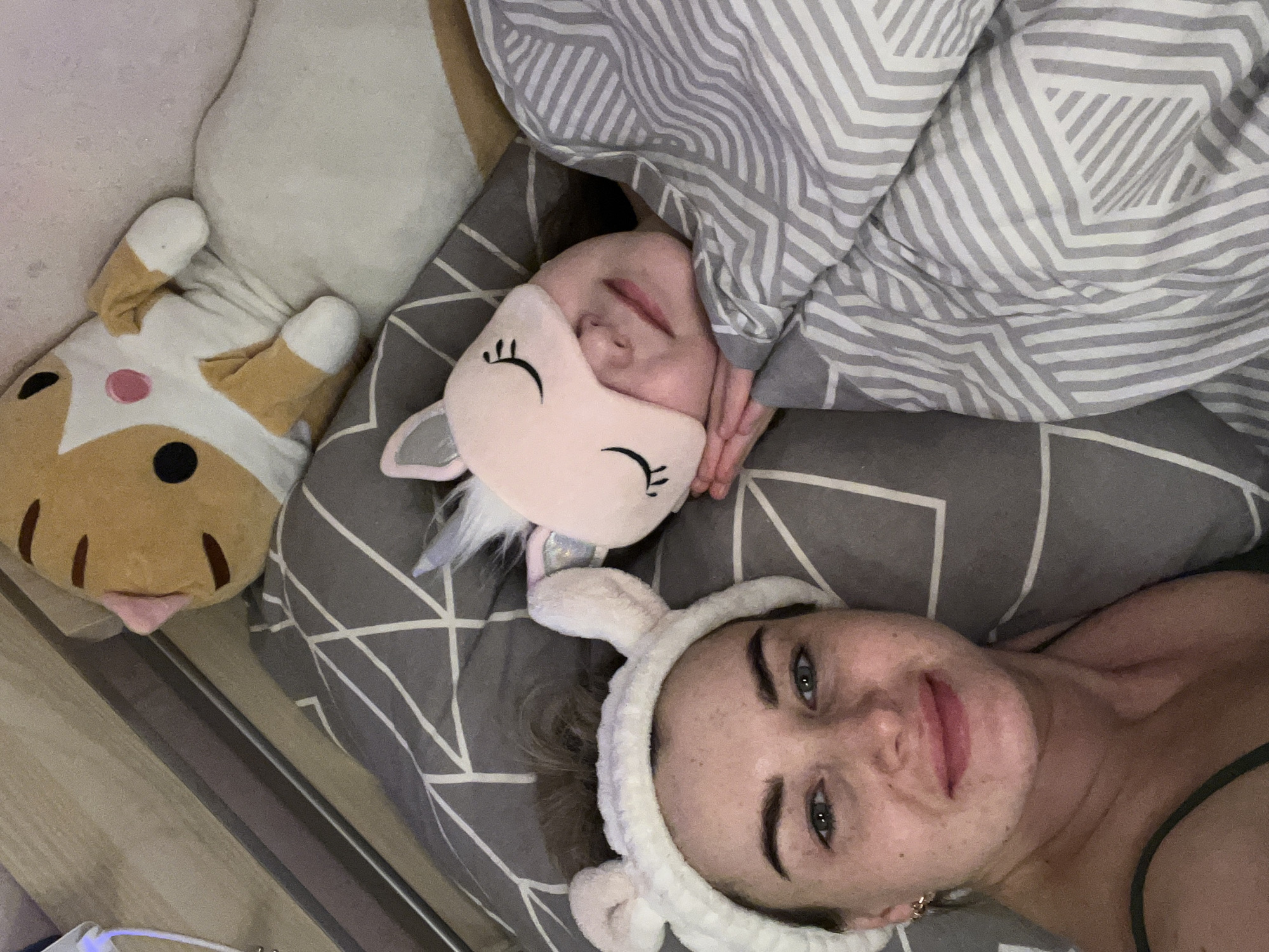 Two individuals, a child and an adult female, are lying in bed with the head of the child resting on a grey pillow with a linear white design and covered by a white-and-grey striped blanket. The child wears a pink unicorn eye mask. The adult female, who is wearing a white headband with small ears, is smiling. To the left of the child is a stuffed animal resembling a cat. The floor is visible on the left edge of the image. The location is not determinable based on the image.