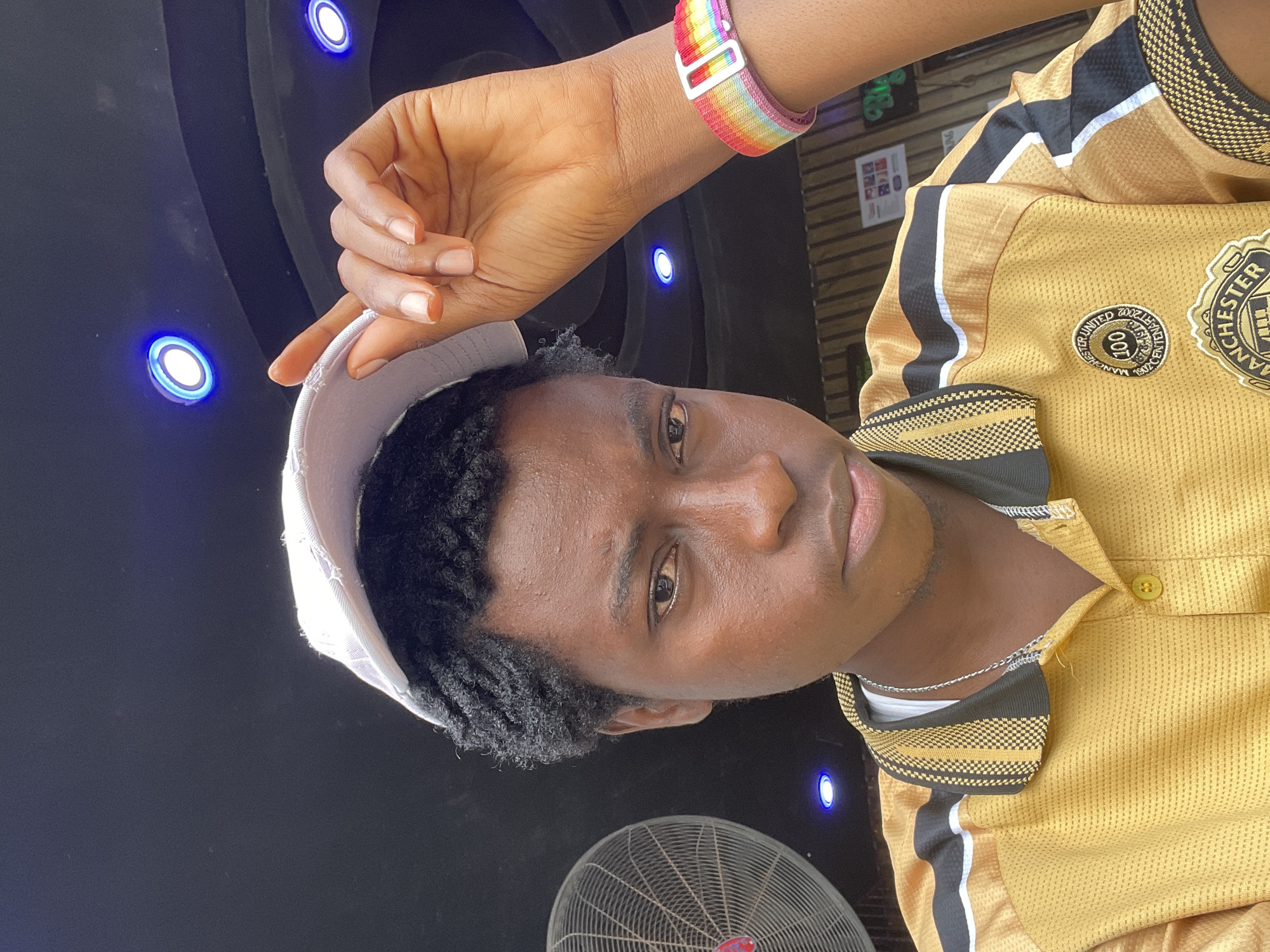 The image shows a young Black person wearing a yellow and black Manchester United jersey. They are also wearing a white cap and a rainbow-colored wristband. The person is looking towards the camera with a neutral expression. The background is dark and appears to be indoors, with some blue lights visible. There is also a fan in the lower part of the image. The overall impression is that of a casual, everyday moment captured in a potentially urban or social setting. The visible text includes "Manchester United 2002 2003" and "100" on a badge on the jersey, as well as some illegible text on flyers in the background. The location is Nkpor, Nigeria.