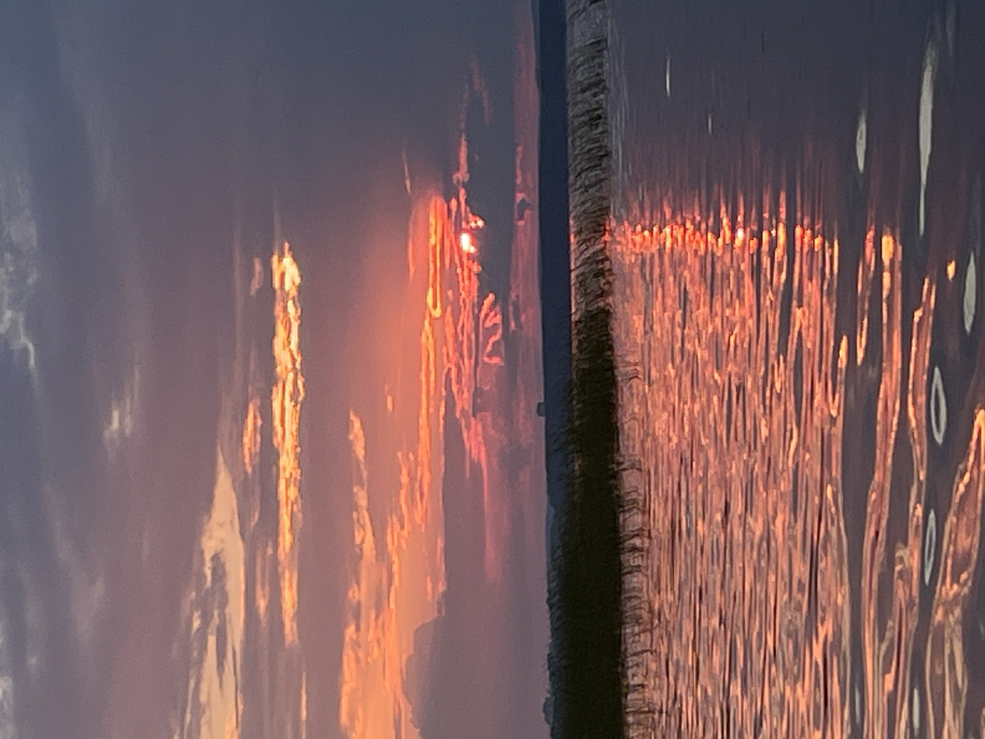 The image depicts a sunset over a body of water, likely the Kama River, near Chaikovsky, Russia. The sky is filled with dramatic clouds, illuminated by the setting sun, creating vibrant hues of orange, pink, and purple. The sun itself is partially obscured by clouds, casting a warm glow on the scene. In the foreground, the water reflects the colors of the sky, creating a shimmering effect. A dark line of vegetation, possibly reeds or tall grass, runs along the horizon, separating the water from the sky. In the distance, a few indistinct structures can be seen on the horizon. The overall impression is one of natural beauty and tranquility.