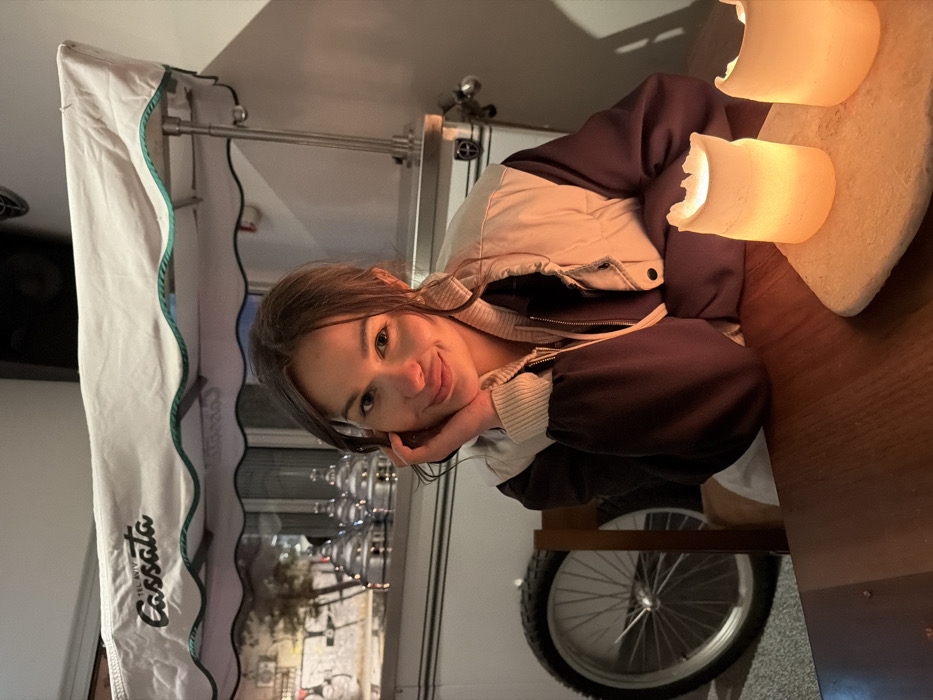 A young adult female is depicted seated at a wooden table in an establishment. She faces the viewer with a slight smile, her right hand resting on her cheek. She wears a light-colored vest over a dark long-sleeved garment. On the table, two illuminated faux candles sit on an irregular light-colored base. Behind her, a mobile cart with a white canopy displaying "TEL AVIV Cassata" in dark script is visible. The cart features multiple reflective containers on its surface and a spoked wheel on its left. Reflections of an outdoor environment, possibly a street, are discernible through the cart's panels. The scene is dimly lit with warm ambient light. The location is specified as Tel-Aviv, Israel, based on the visible signage.
