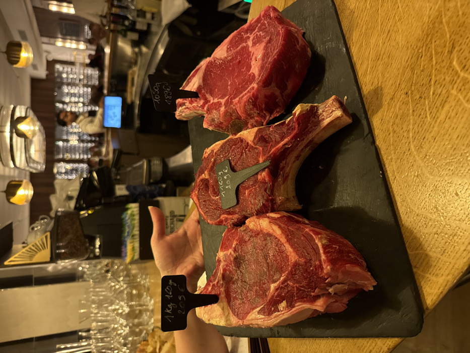An interior shot depicts three raw, bone-in cuts of red meat, likely beef ribeye or côte de boeuf, presented on a rectangular black slate board, resting on a light brown wooden table. Each cut features a black spade-shaped marker indicating its weight and price in Euros. The leftmost cut displays "1 kg 160g / 205 €," the middle cut "1.160 g / 205 €," and the rightmost cut "1050 g / 189 €." A light-skinned hand is visible on the upper left, positioned near the display. In the background, an upscale restaurant or steakhouse environment is visible, consistent with a location in Paris, France. Clear wine glasses are stacked on the left, next to bread. On the right, a dark-haired male individual, wearing a light shirt and dark apron, stands behind a bar counter with a blue-lit monitor and various bottles, including wine, visible. The lighting is warm and ambient, featuring decorative ceiling fixtures.