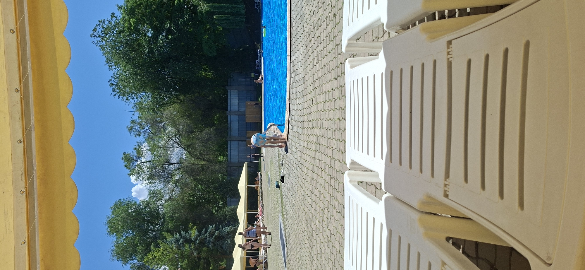 A poolside scene under a yellow awning on a bright, sunny day. In the foreground, two white lounge chairs are partially visible, suggesting an outdoor recreation area. Beyond the chairs, a paved area with patterned stone leads to a bright blue swimming pool. Several people are present: two women in swimsuits are walking on the pavement to the left, under yellow canopies that provide shade over lounge chairs. A child in blue swim trunks is bending down at the edge of the pool, holding onto the railing. Another person is sitting on a lounge chair by the pool. Trees and a fence line the background. The image appears to be taken in Almaty, Kazakhstan.