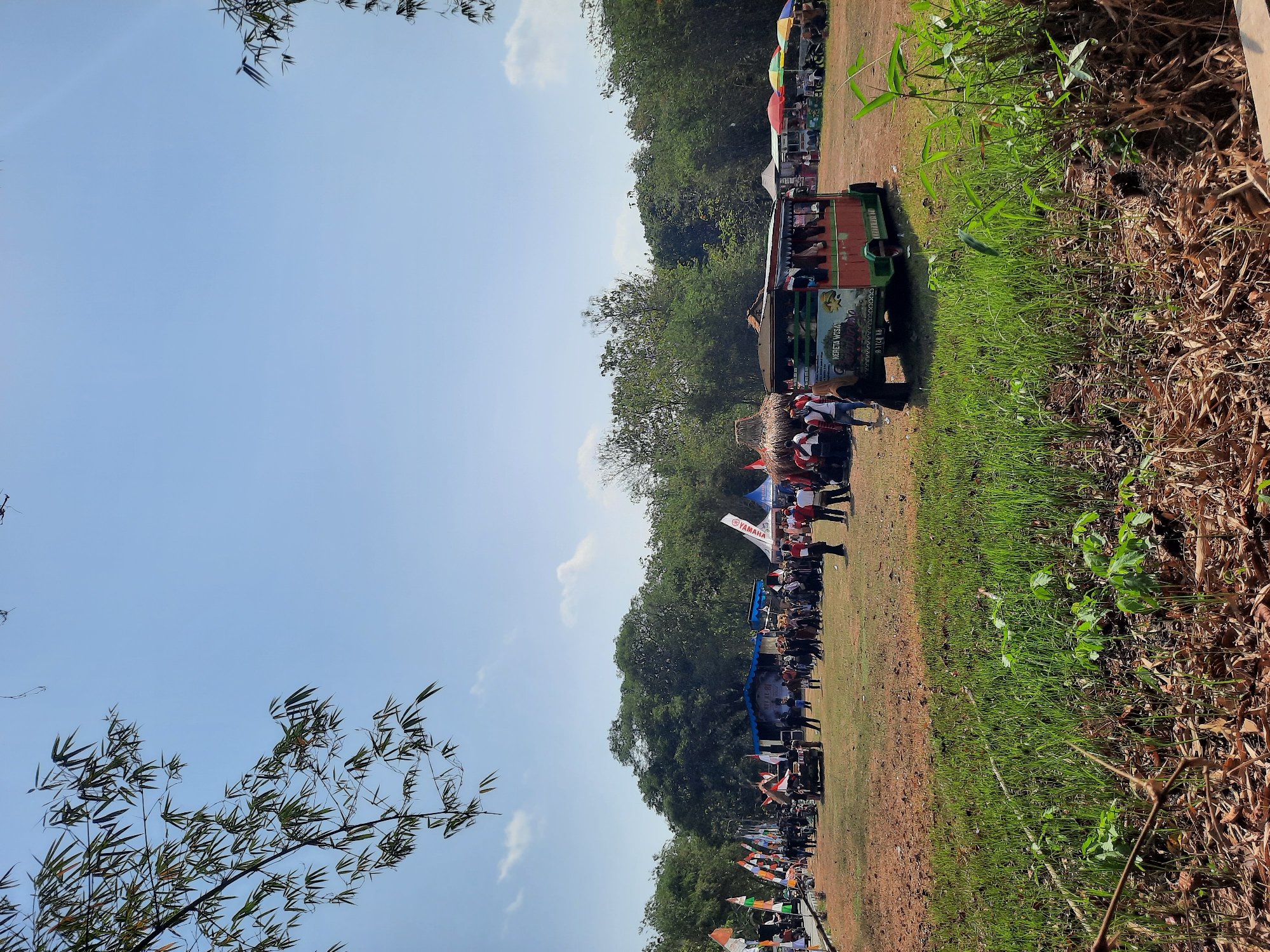This is a full, eye-level outdoor shot of a community event. The event appears to be taking place in Lugu, Indonesia, indicated by the presence of Indonesian flags and a sign on the "Kereta Wisata" vehicle that translates to "Tourist Train." The scene is set in a field with a dry, dusty ground in the foreground, transitioning to green grass and dense trees in the background. A bright blue sky with scattered clouds dominates the upper portion of the image.
In the mid-ground, a crowd of people is gathered around various stalls and a small stage. Several flags, including the Indonesian flag, are planted in the ground. A distinctive "Kereta Wisata" vehicle, painted red and green, is parked on the right side of the frame, with several people inside and around it. In front of the vehicle, a woman in a headscarf is walking away from the camera. Numerous stalls with colorful umbrellas are set up, suggesting a market or food vendors. The presence of motorcycles indicates attendees may have arrived by personal transport.
In the foreground, lush green grass and dry leaves cover the ground, with some thin branches and debris scattered across it. Bamboo branches are visible in the upper left and right corners of the image, framing the scene and suggesting the location is near a wooded area. The overall atmosphere is lively, with many people attending what appears to be a local festival or gathering.