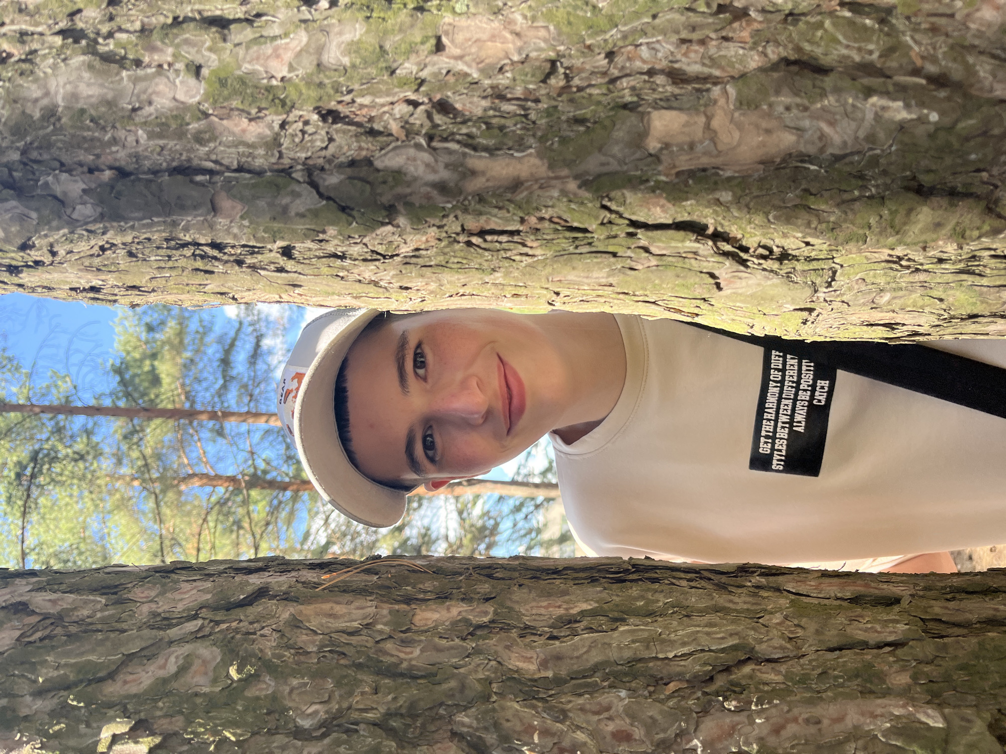 In this medium-shot, outdoor portrait, a young person with short dark hair and a light-colored baseball cap smiles at the camera, positioned between two large tree trunks. The person wears a light-colored crewneck sweatshirt with a black patch on the chest featuring white text. The background consists of a forest scene with tall pine trees under a bright blue sky with scattered clouds. The lighting suggests it is daytime. The location is the city of Chaikovsky, Russia.