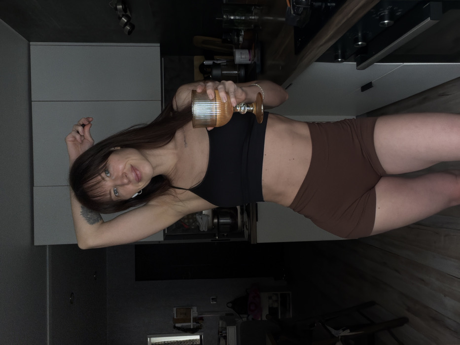 The image depicts a female individual in a kitchen located in Perm, Russia. The subject, possessing brown hair with bangs, is clad in a black spaghetti strap crop top and brown high-waisted shorts. White wireless earbuds are visible in both ears. Tattoos are present on her upper left arm and faintly on her upper chest. She is captured in a pose with her left arm raised, hand behind her head, and her right hand extended forward, holding a ribbed glass goblet containing an amber-colored liquid. Her facial expression shows a faint smile. The kitchen environment includes white upper cabinets with dark handles, a dark countertop, and dark wooden floorboards. A stovetop and an oven are visible on the right side of the countertop. In the background to the left, a window, a dark tall cabinet, and a table area with miscellaneous objects are discernible. The scene is illuminated by dim, ambient light.