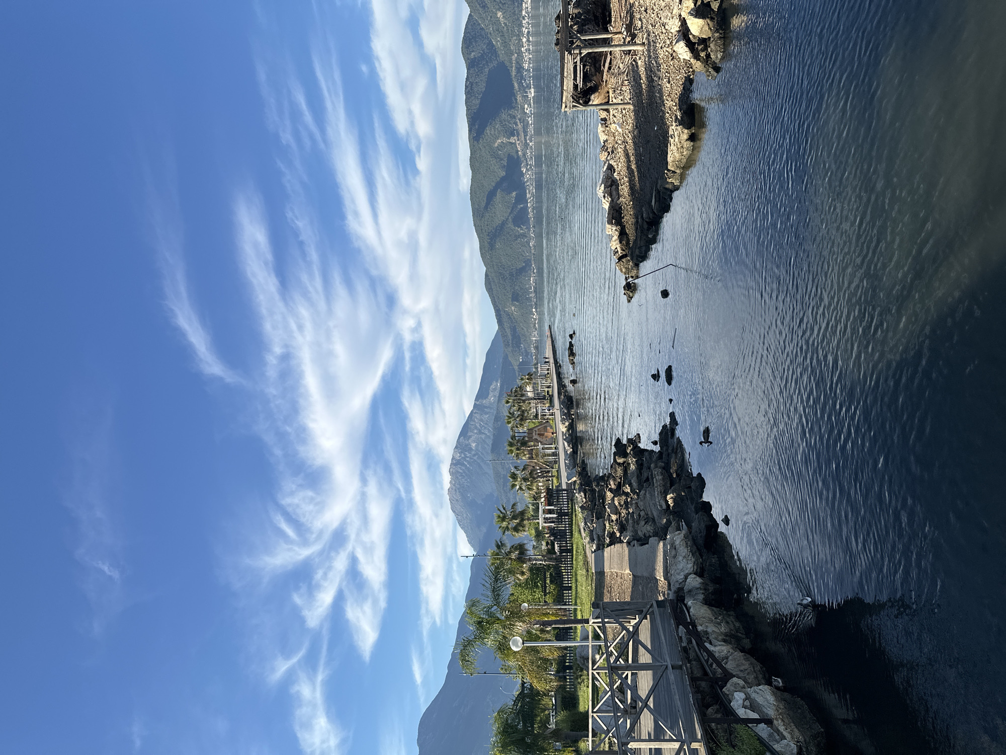 The image shows a park in Fethiye, Turkey along the sea. Tall mountains loom in the background under a blue sky with wispy clouds. A boardwalk with a wooden railing edges the park, leading to a stone path. Palm trees and lampposts line the park. The sea is calm with rocks along the edge. A small structure sits on the rocks jutting into the water.
