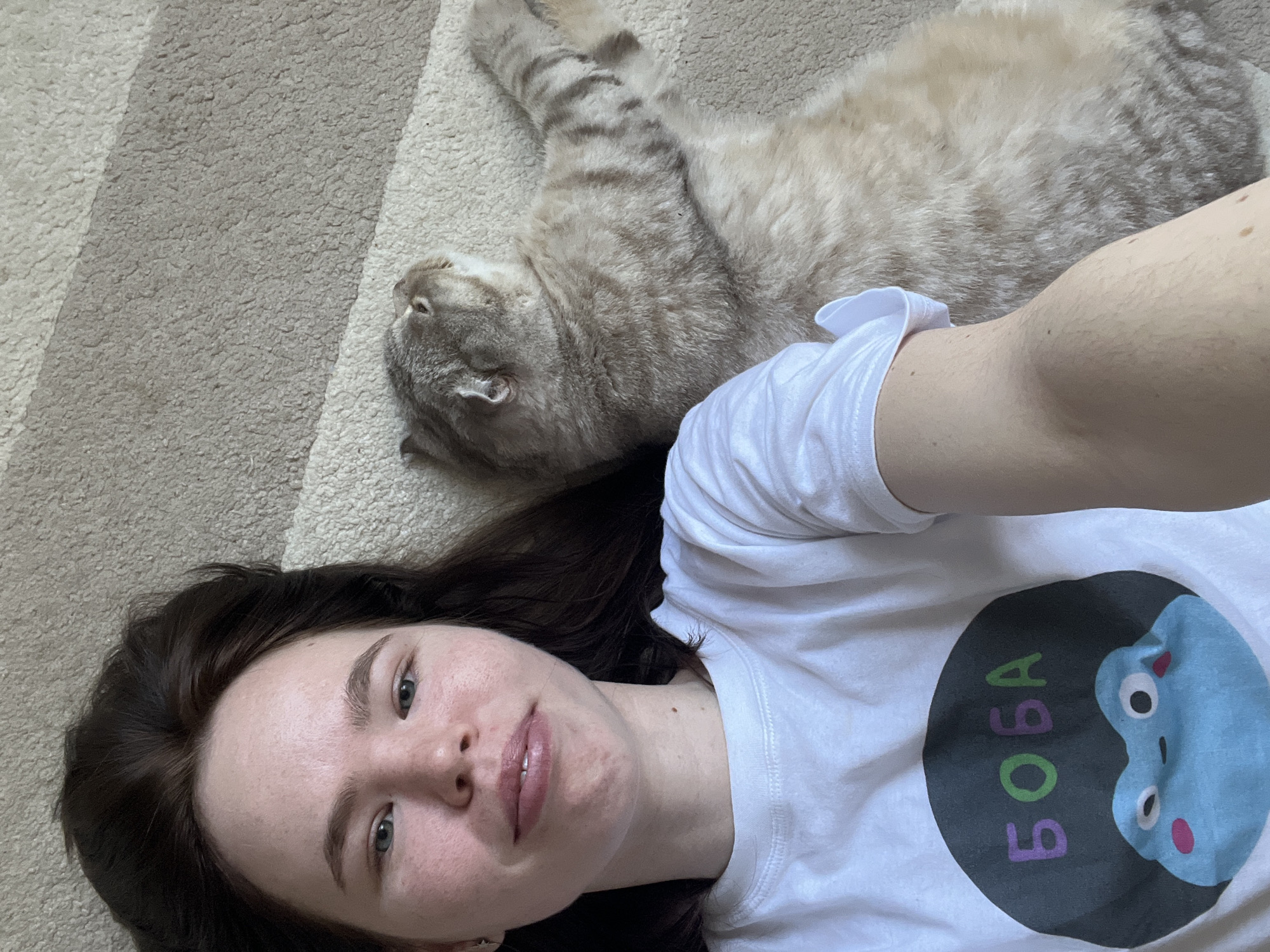 A young woman takes a selfie while lying on her back on a striped beige carpet, with a light-colored tabby cat resting beside her. The scene is set indoors in Chaikovsky, Russia, during what appears to be daytime, indicated by the bright, even lighting.
The woman, with dark hair, looks directly at the camera with a calm, slightly smiling expression. She is wearing a white short-sleeved t-shirt featuring a large circular graphic. This graphic displays a light blue, frog-like character with pink cheeks and white eyes. Next to the character, the word "БОБА" is written vertically in green and purple Cyrillic letters. The cat is also lying on its back, appearing relaxed with its head turned away from the camera and its paws somewhat splayed. The carpet has a textured, striped pattern in varying shades of beige. The overall mood of the image is casual and domestic.