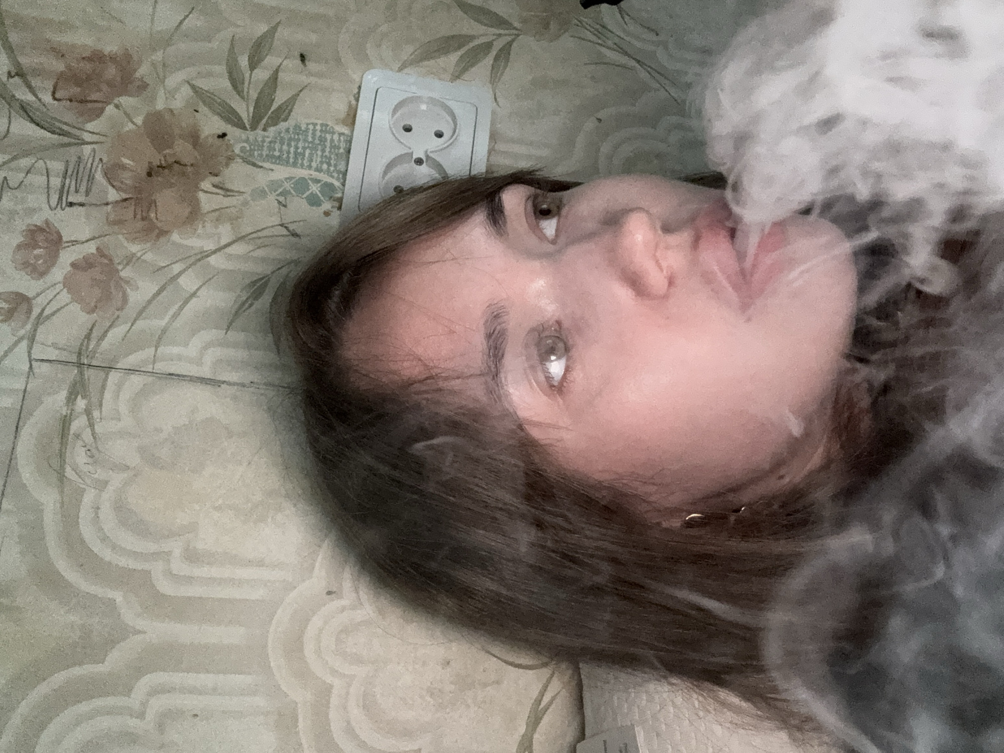 A young woman with brown hair lies on a light-colored patterned surface, possibly a floor or wall, in a residential setting. Her head is near a white electrical outlet. Smoke, or possibly vapor, is exhaling from her mouth. It cannot be determined from the image if this image was taken in the city of Chaikovsky, Russia.