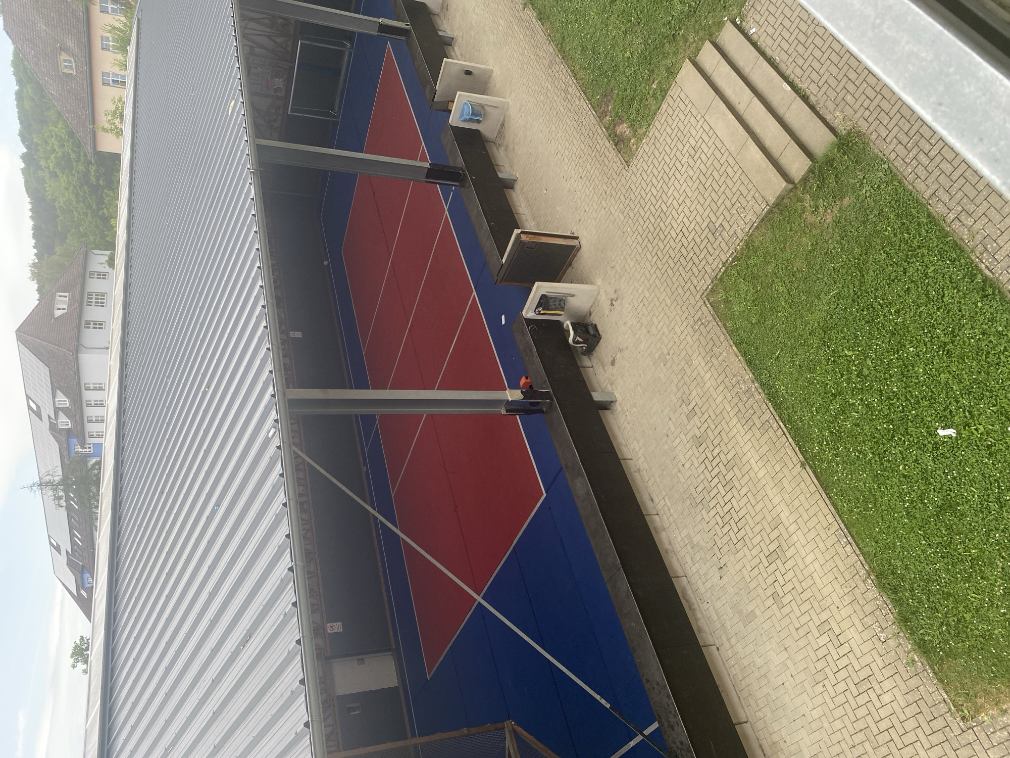 The image shows an outdoor sports court, likely for basketball or volleyball, located in Göttingen, Germany. The court has a red playing surface bordered by a blue outer area. A metal roof supported by pillars covers the court. Adjacent to the court is a paved walkway with several concrete structures that appear to be benches or trash receptacles. Green grass and trees are visible surrounding the paved area and in the background, with buildings and a wooded hillside in the distance. The sky is overcast.