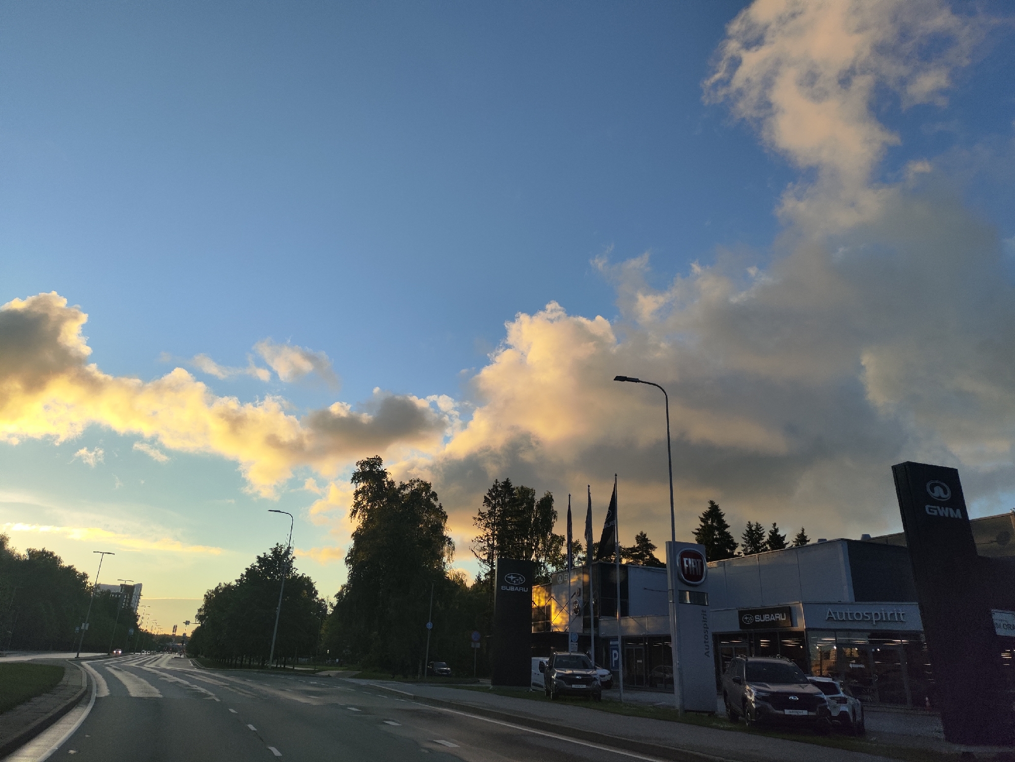 The image captures a street view in Tallinn, Estonia, during what appears to be sunset or early evening. A multi-lane road curves to the left, with a grassy median on the left side. On the right side of the road, a car dealership is visible, displaying prominent signage for "FIAT", "SUBARU", and "Autospirit". A large black pylon with "GWM" logo is also present. Several cars are parked in front of the dealership, including a dark SUV and a lighter-colored sedan. Trees line the right side of the road and the dealership lot. The sky above is a mix of clear blue with scattered clouds that are illuminated with a warm, golden light, indicating the time of day. Streetlights are positioned along the road. The overall scene depicts a commercial area on the outskirts of the city as daylight fades.