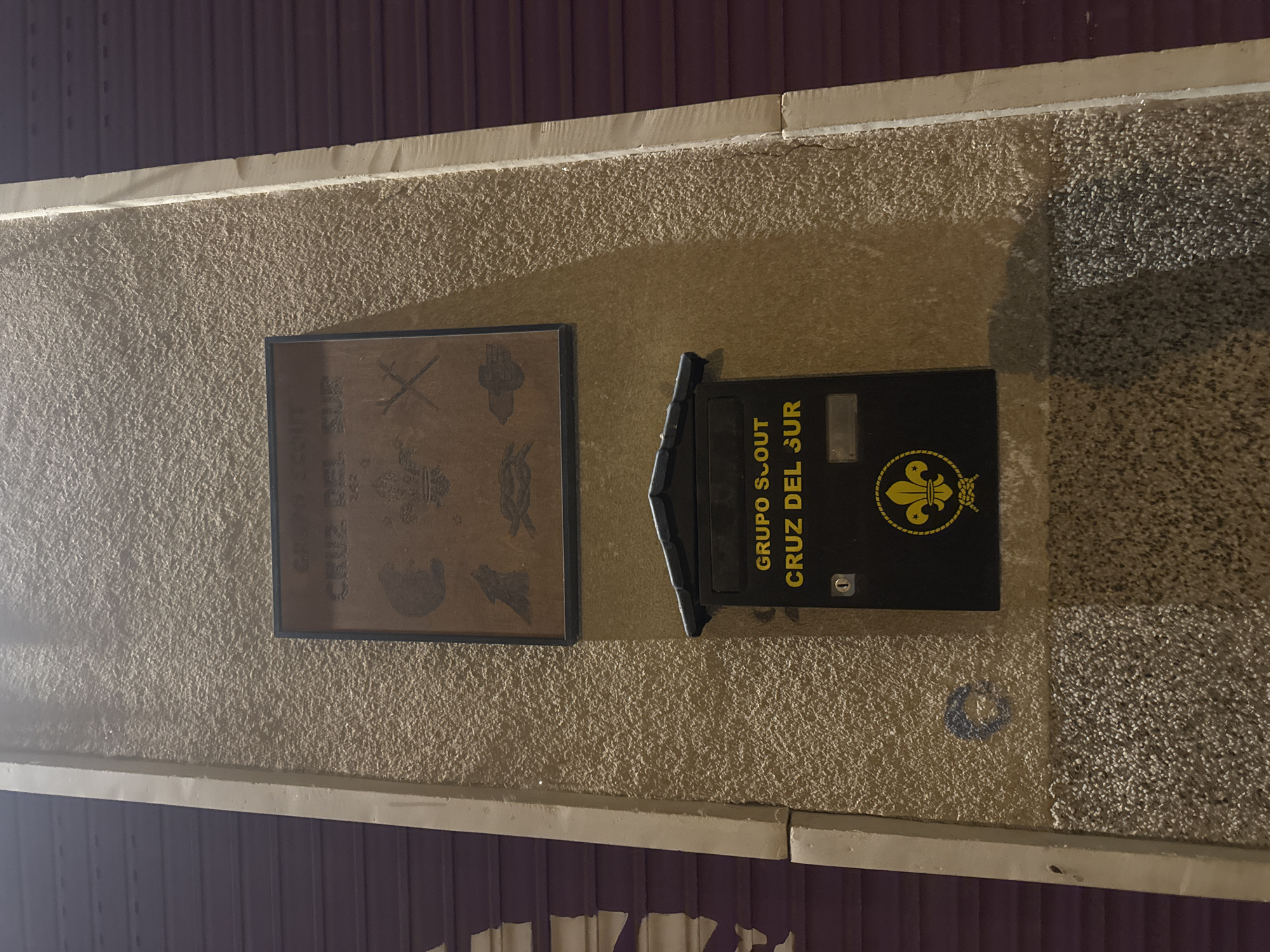 The image shows a close-up view of a textured, light-brown wall with a dark purple corrugated metal shutter to the left and right. Mounted on the wall are two distinct items. The upper item is a square plaque with a dark border, featuring an embossed design. The text at the top of the plaque reads "GRUPO SCOUT" and below that "CRUZ DEL SUR". The plaque contains several embossed emblems, including what appears to be a wolf cub, a fleur-de-lis, and other scout-related symbols.
Below the plaque is a black mailbox with a peaked roof. The mailbox also has yellow text that reads "GRUPO SCOUT" at the top and "CRUZ DEL SUR" below it. Centered on the front of the mailbox is a large, yellow fleur-de-lis emblem, a common symbol for the Scout movement. To the left of the fleur-de-lis is a small keyhole. The wall surface has a rough, stucco-like texture. There is a circular blue marking on the wall below and to the left of the mailbox. The lighting suggests it is nighttime, with light illuminating the wall and objects. The context provided indicates this is located in the city of Murcia, Spain.