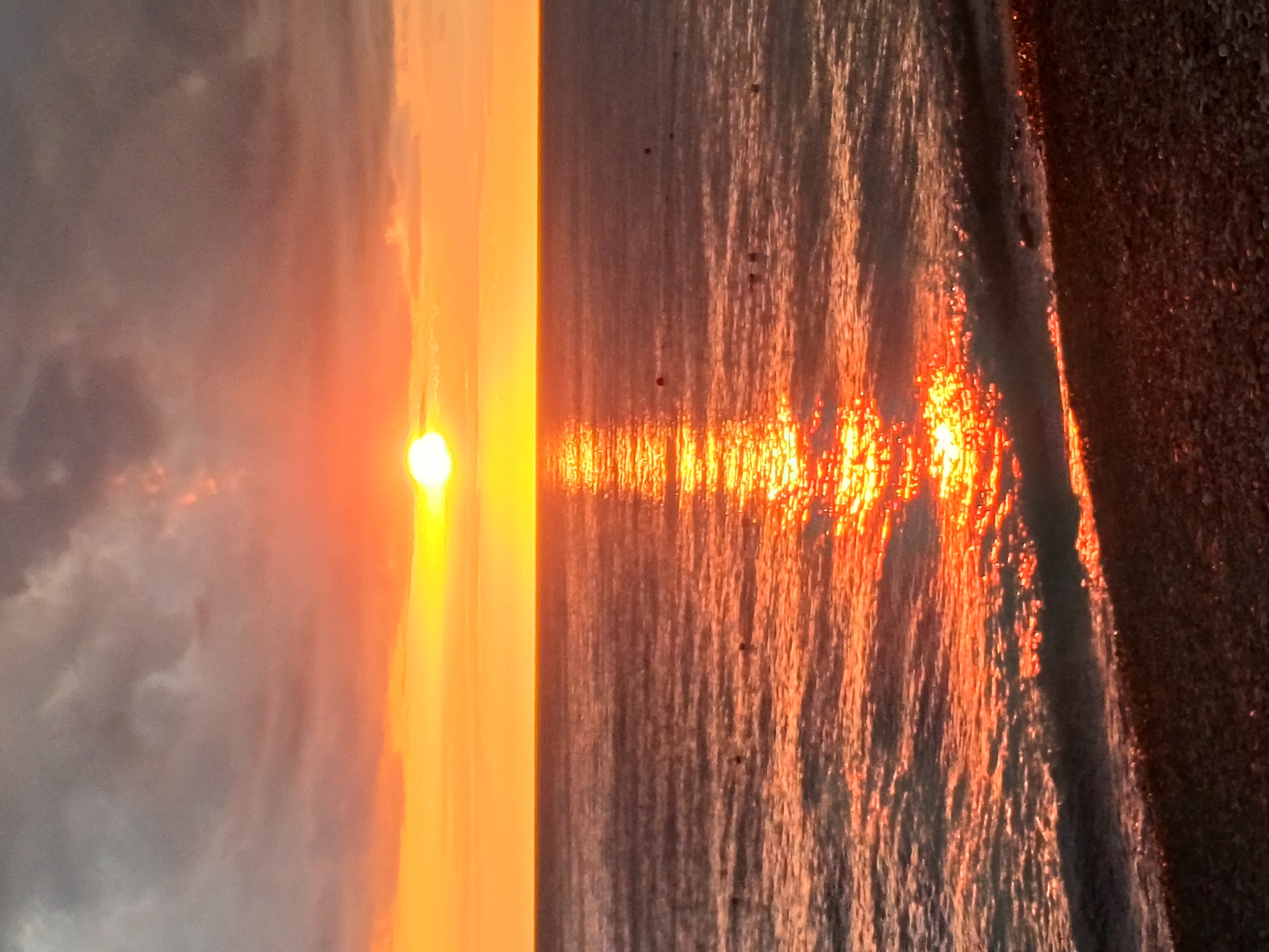 This is a vertical, eye-level, medium shot of a sunset over the Black Sea in Sochi, Russia. The sun is a bright, glowing orb near the horizon, casting a golden reflection across the water. The sky is a mix of orange, yellow, and gray clouds. The water is calm with small waves lapping at a pebble beach in the foreground. There are a few buoys visible in the water.