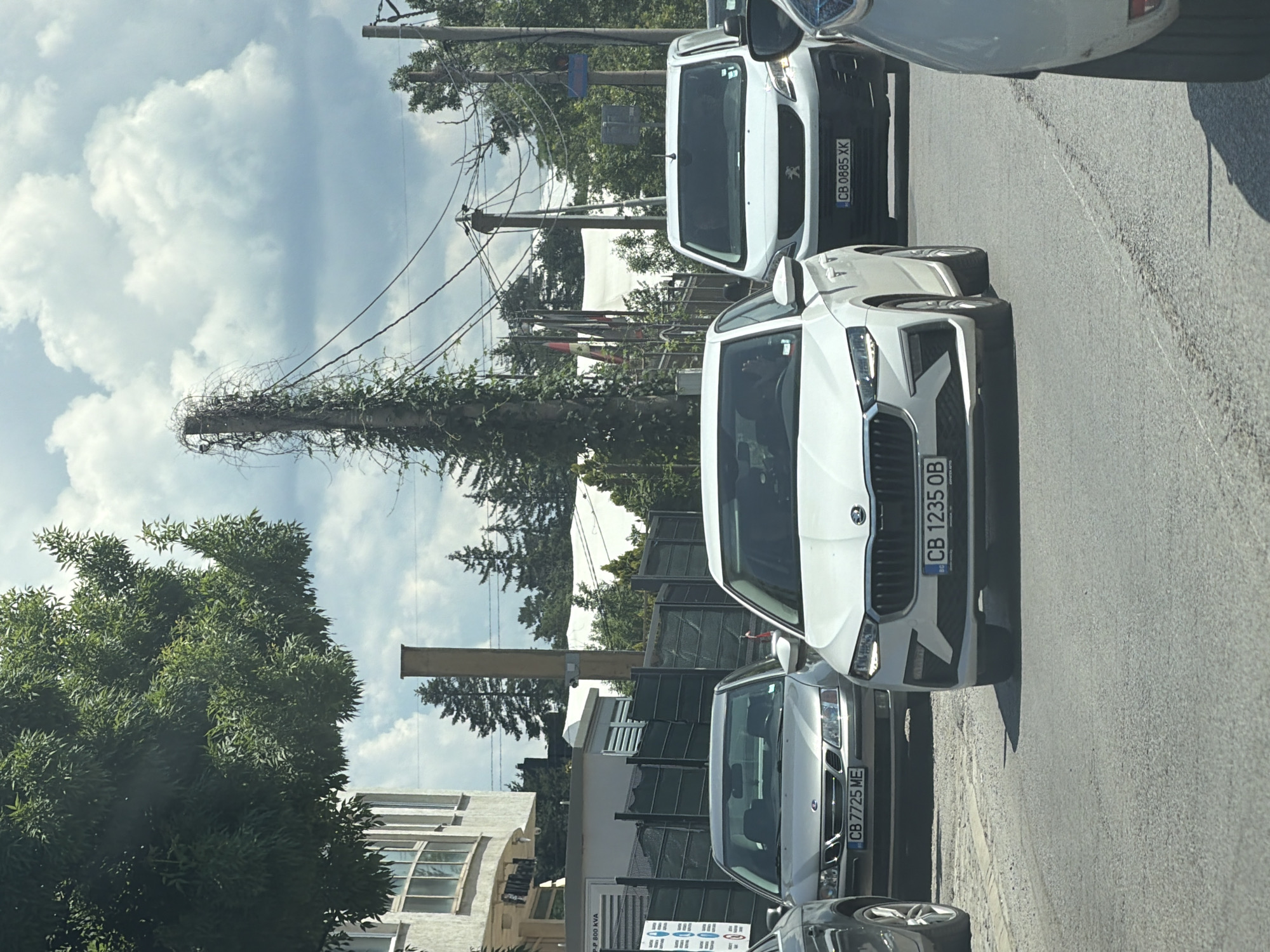 In Sofia, Bulgaria, a traffic jam is captured on a sunny day. The image focuses on several cars: a white Skoda, a silver Saab, a white van, and another white car. The Skoda in the foreground displays the license plate "CB 1235 OB". Behind it, the Saab has a license plate reading "CB 7725 ME", and the van shows "CB 0885 XK". Trees and buildings line the street, with power lines overhead. The sky is partly cloudy, indicating fair weather. The scene depicts a typical urban traffic situation.