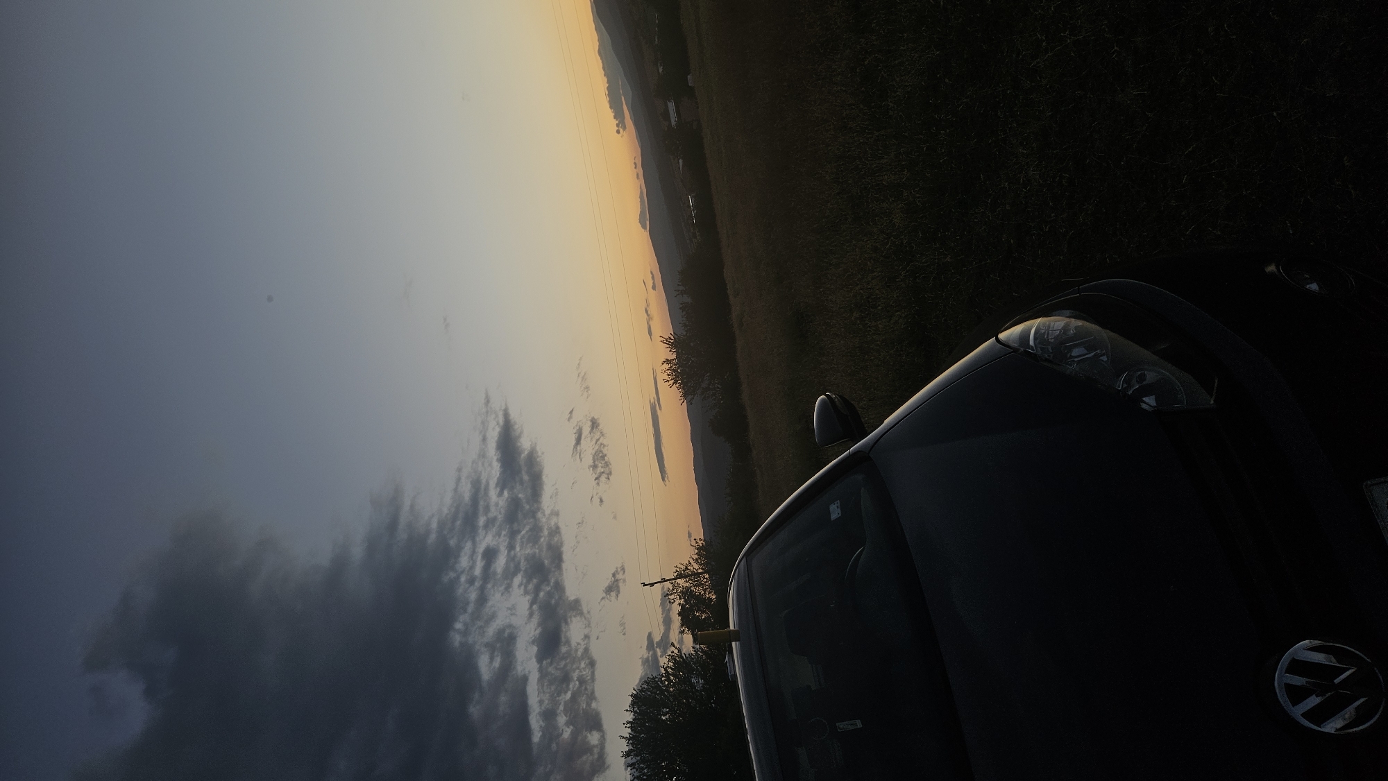 A dark Volkswagen car is parked in a grassy field with trees and hills in the background. The sky is a gradient of dark gray clouds on the left to a pale orange sunset on the right. The overall scene is dimly lit, suggesting it is either early morning or late evening.