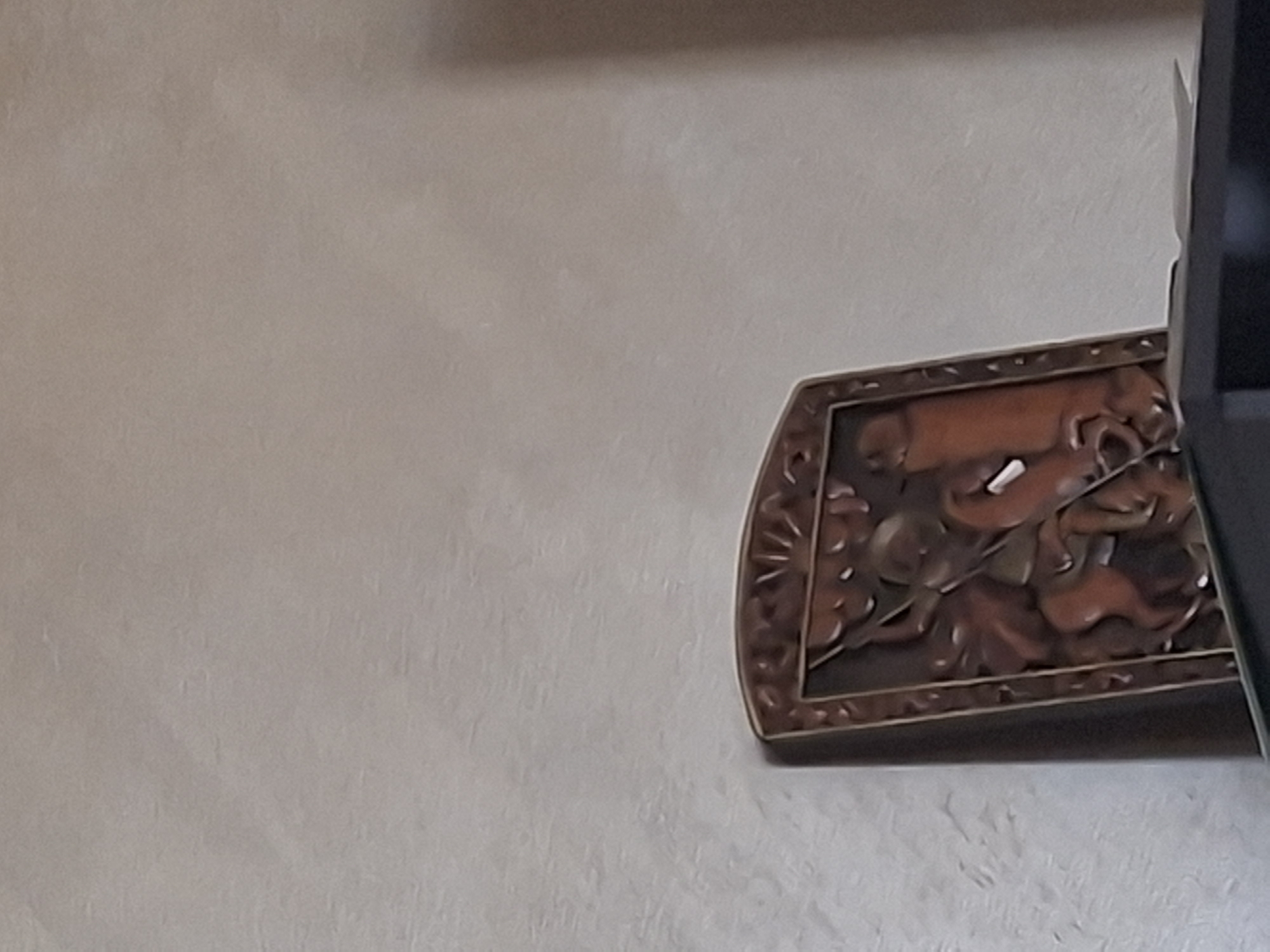 A carved wooden plaque depicting a warrior on horseback is displayed on a shelf. The plaque is rectangular with a decorative border. The carving shows a figure in armor with a sword, facing a dragon or serpent-like creature. The warrior is mounted on a horse that is rearing. The background of the plaque is textured. The plaque is positioned on the right side of a dark shelf, with a portion of the shelf and the wall behind it visible. The wall has a light, textured finish. The location in Aksakovo, Bulgaria is not discernible from the image.