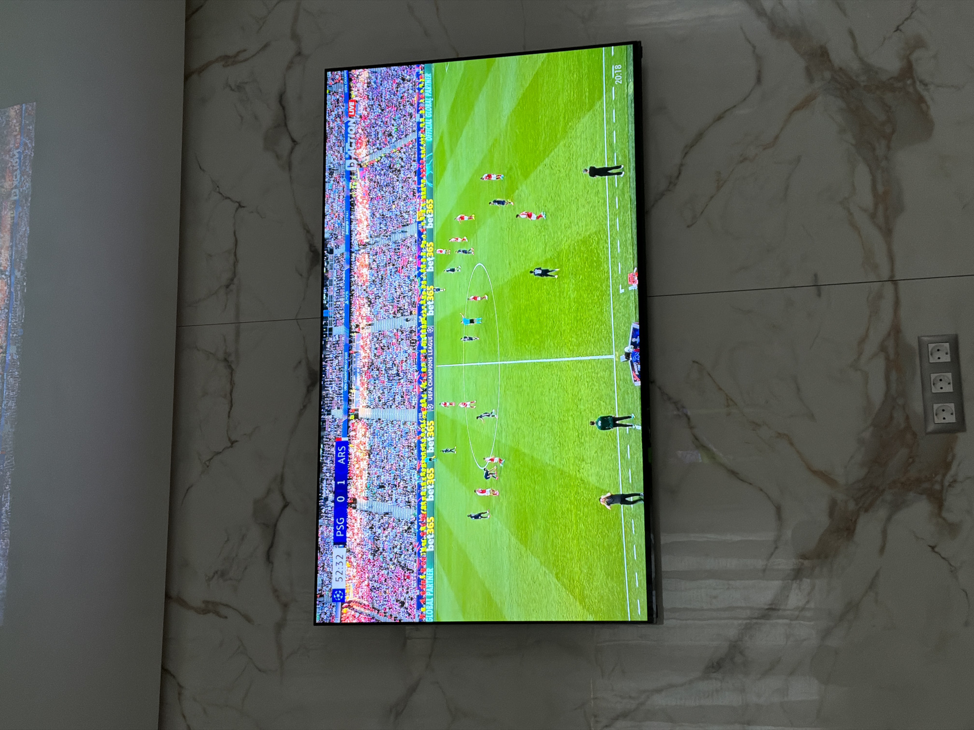 The image shows a television mounted on a marble-patterned wall. On the screen is a broadcast of a soccer match, displaying the score as PSG 0-1 ARS. The match appears to be in the 52nd minute, as indicated by "52:32" at the lower left of the screen. Logos for "bet365" and "UEFA CHAMPIONS LEAGUE" are visible, along with the tagline "ACTION LIVE" and the time "20:18". To the right of the TV, there is an electrical outlet with three sockets. There is no location visible in the image.