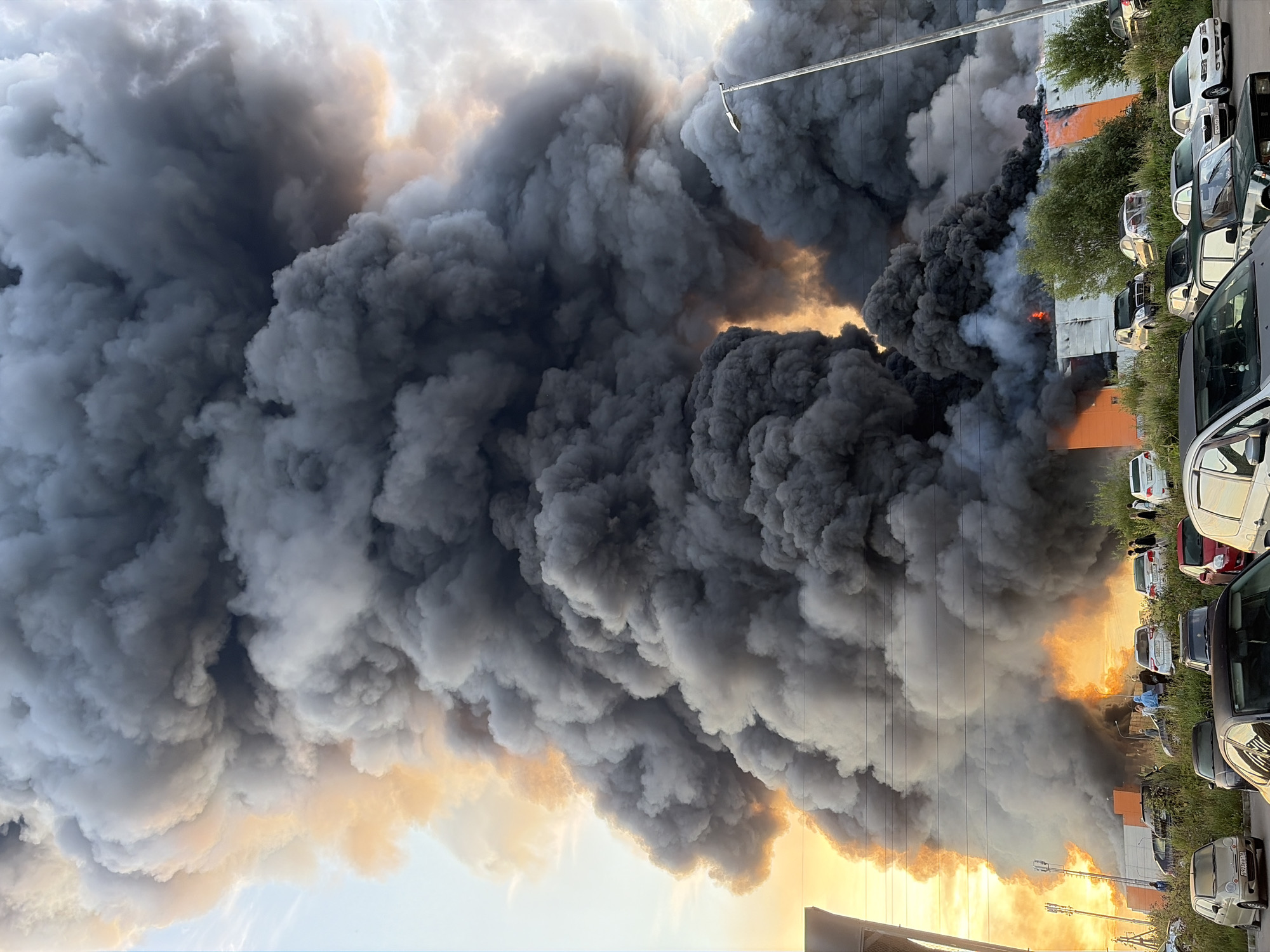 In Nikolskoye, Russia, a massive fire engulfs a building, sending a colossal plume of thick black smoke into the sky. The smoke billows upwards and outwards, dominating the frame and obscuring much of the background. The setting sun casts a warm, orange glow through the smoke on the left side of the image, contrasting with the darker, more ominous smoke on the right. In the foreground, a parking lot is filled with various cars, some parked closely together, while others are more spread out. A few people are visible near the cars, observing the scene. A street lamp stands tall on the right side of the image, partially obscured by the smoke. The building in the background, partially visible beneath the smoke, has an orange and white facade. The scene suggests a dramatic and potentially dangerous event.
