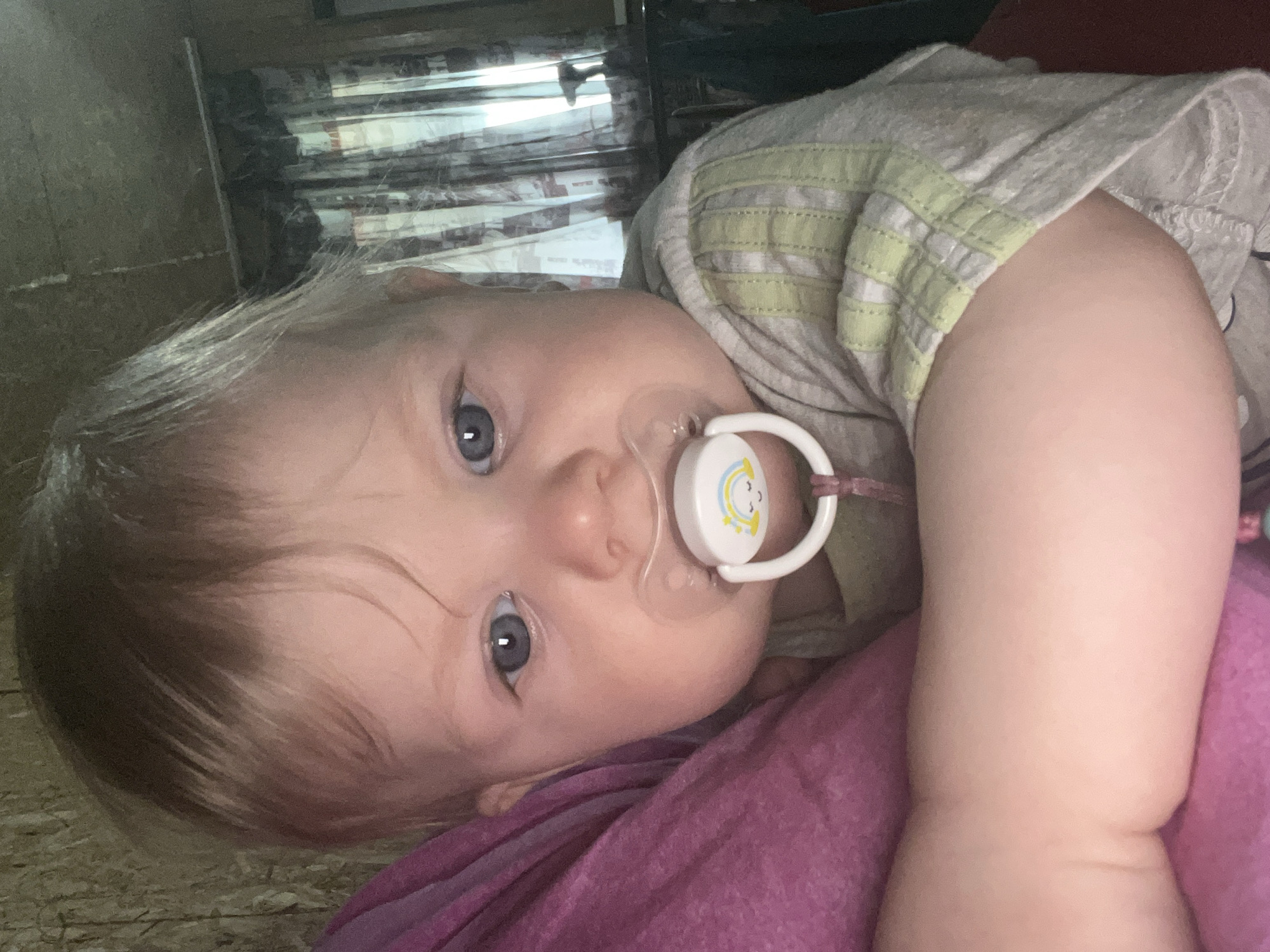 A baby with light blond hair and blue eyes is lying on a purple surface with a pacifier in its mouth. The baby is wearing a light-colored shirt with a yellow and white striped collar. A window with sheer curtains is visible in the background. The location, Vesyoloye, Russia, is not verifiable from the image.